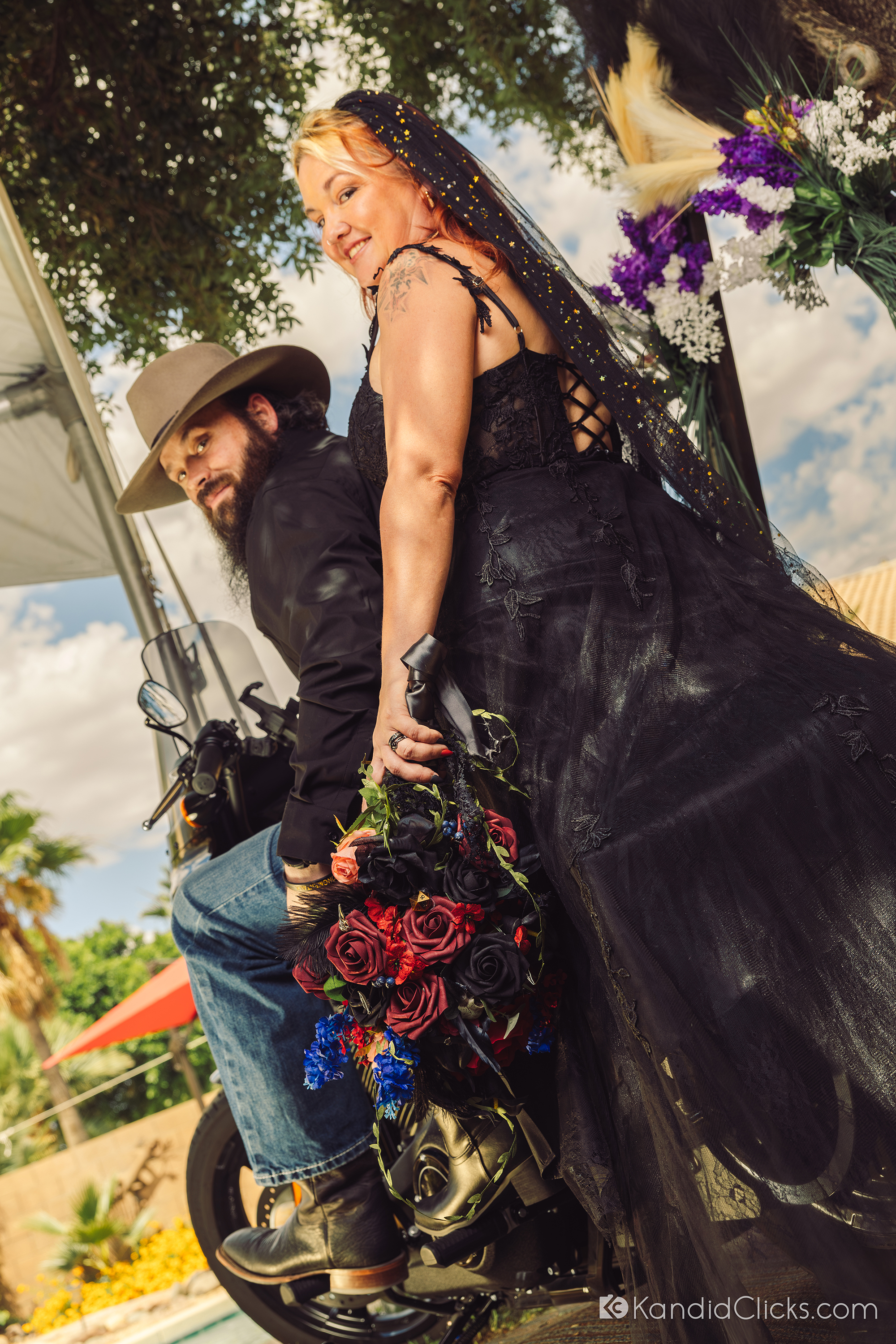 Alternative bride and groom pose confidently on a motorcycle in their wedding attire.