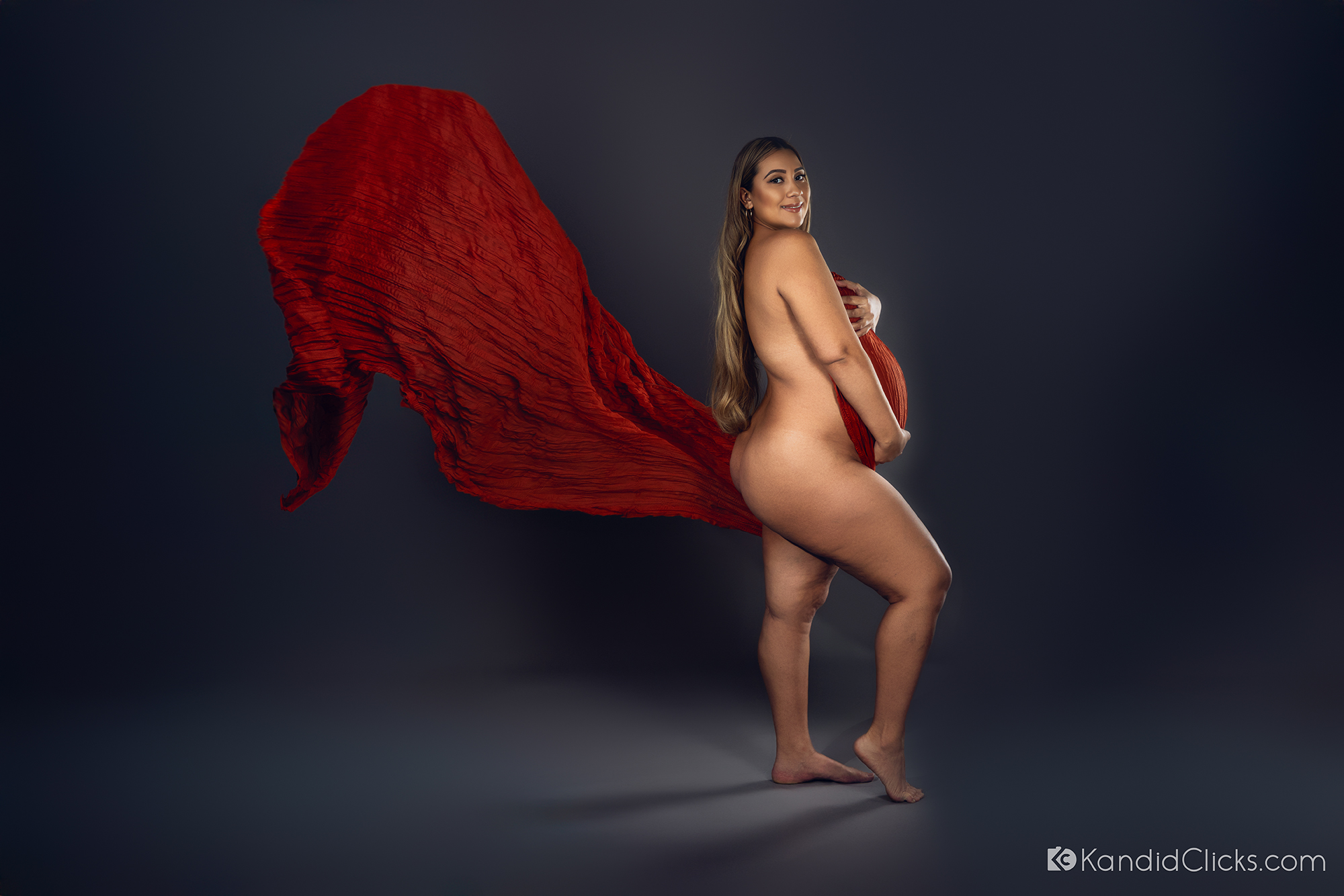 Pregnant woman posing confidently in studio with flowing red fabric behind her