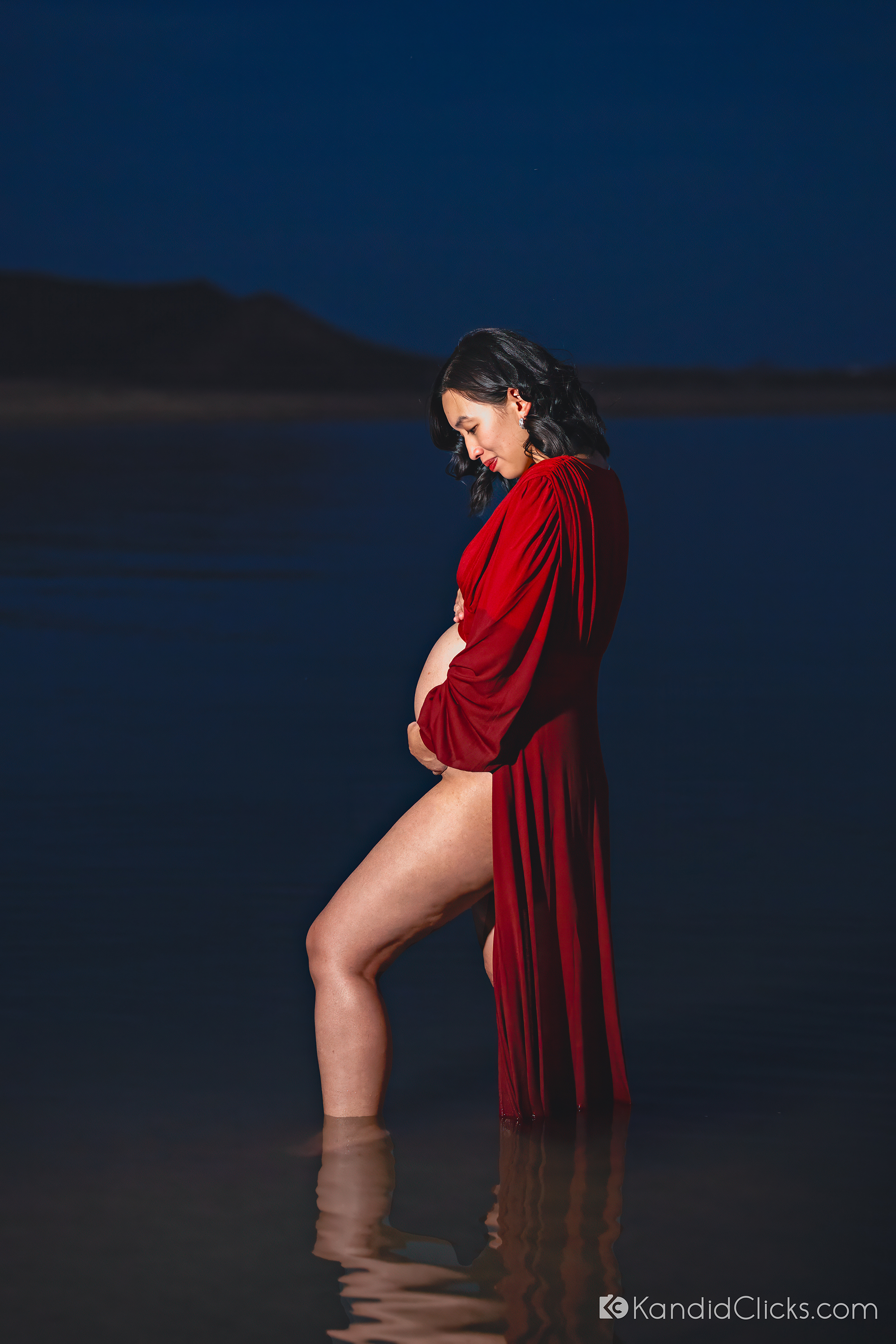 Pregnant woman in red dress standing in water during an evening maternity photoshoot