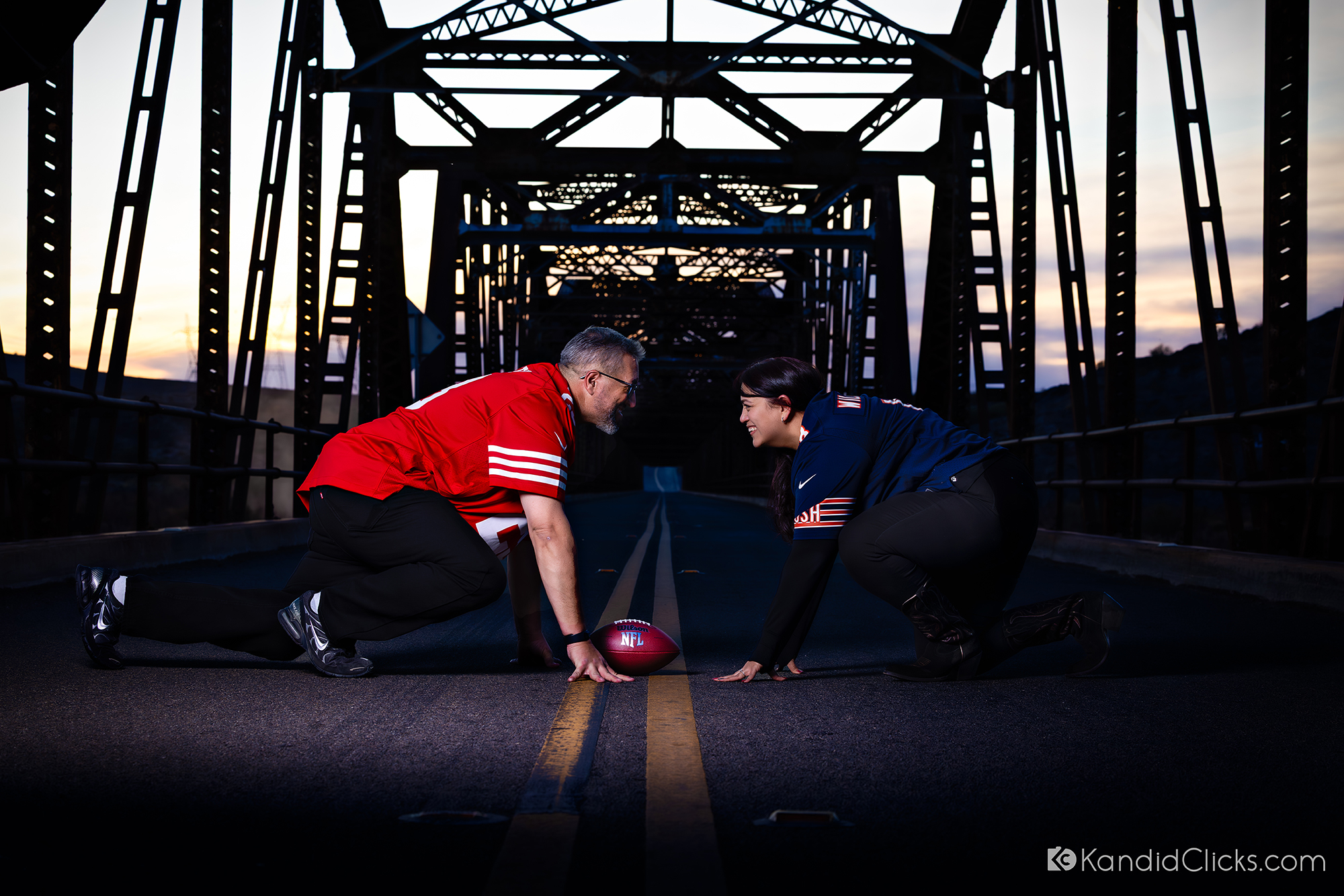 Couple in football jerseys posing on a bridge in a play formation at dusk.