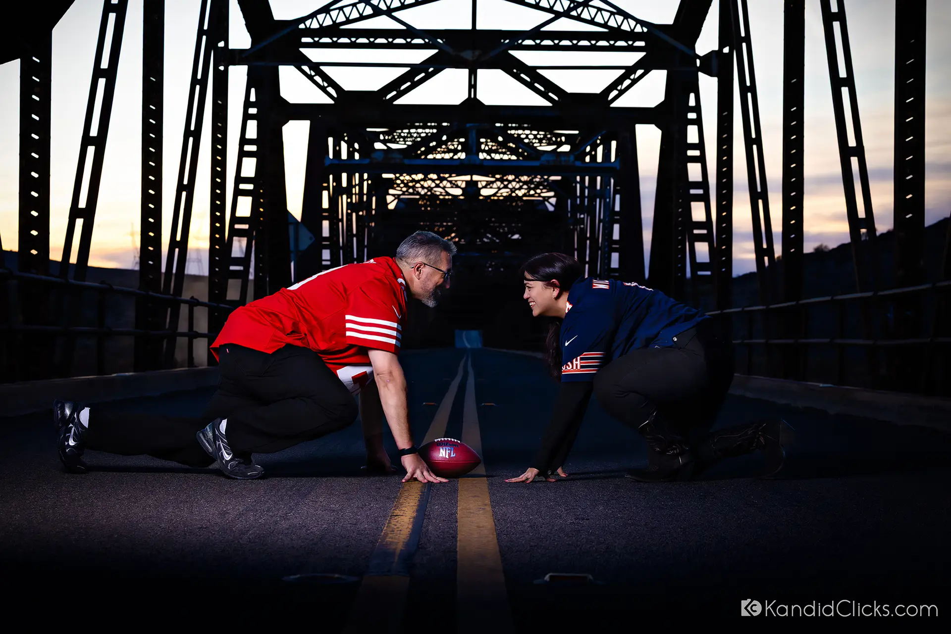 Couple in football jerseys posing on a bridge in a play formation at dusk.
