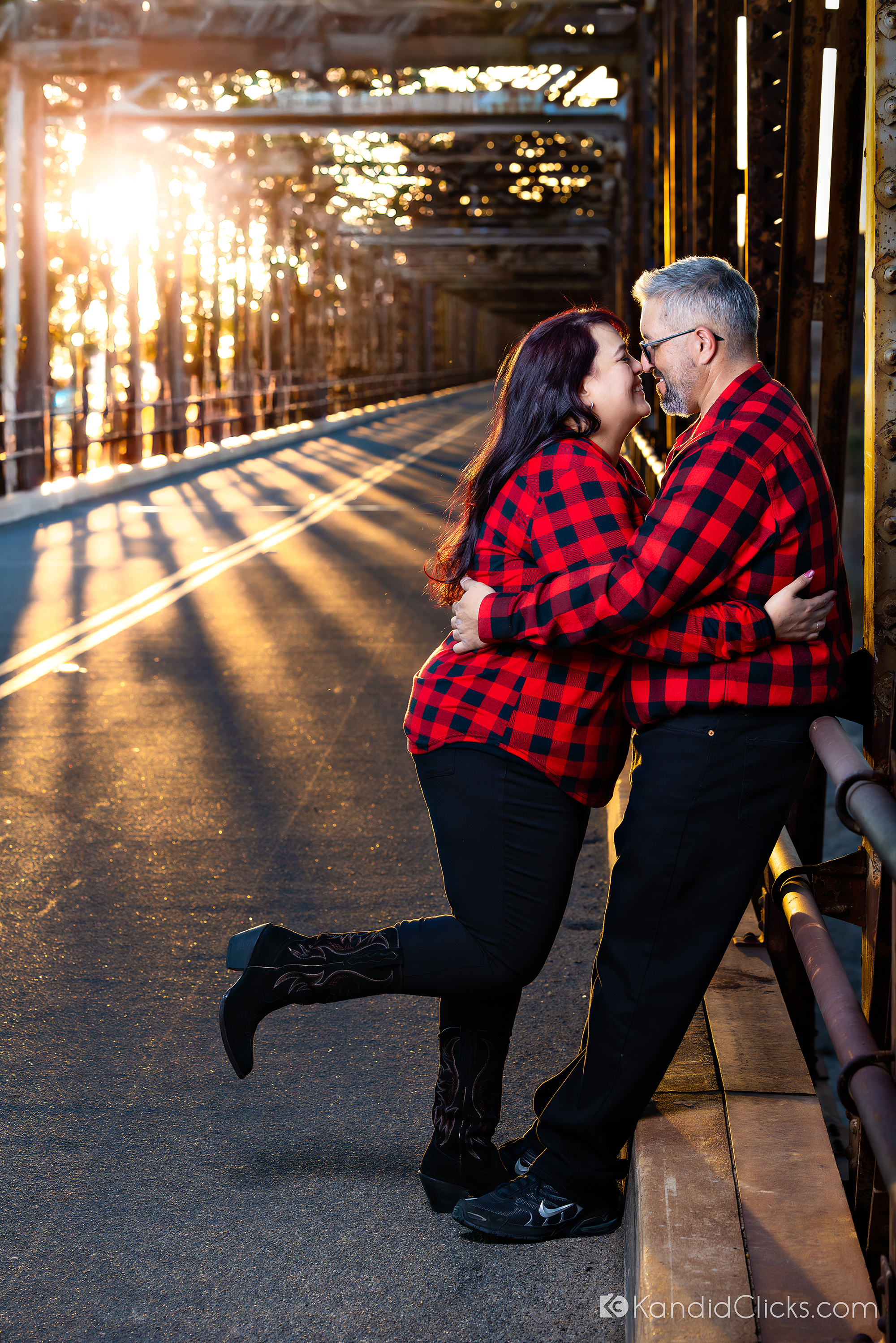 Couple in matching red plaid shirts sharing a romantic moment on a sunlit bridge.