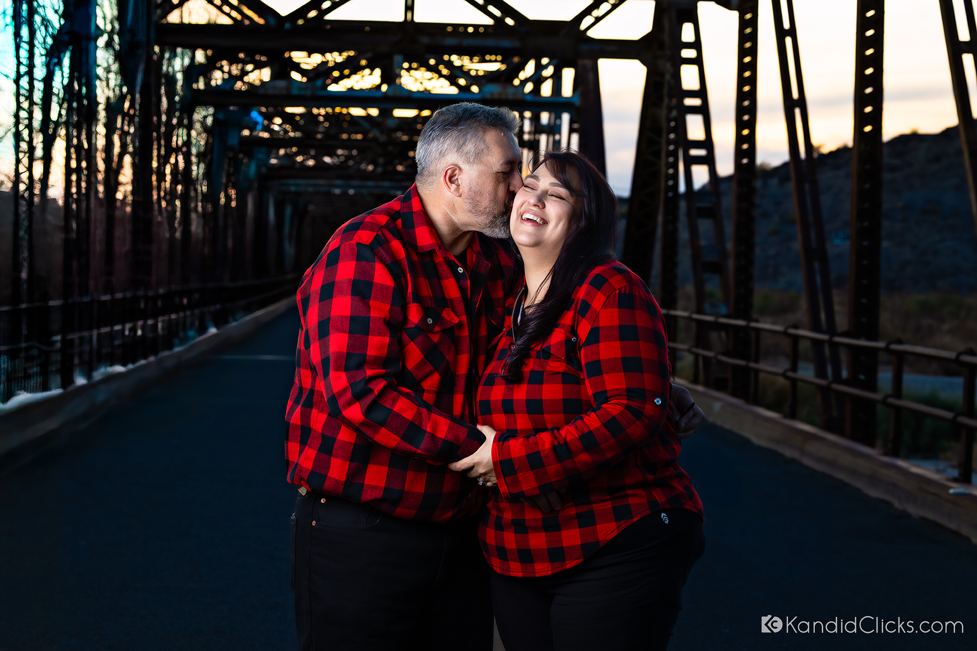 Smiling couple in matching red plaid shirts sharing a tender moment on a rustic bridge at dusk.