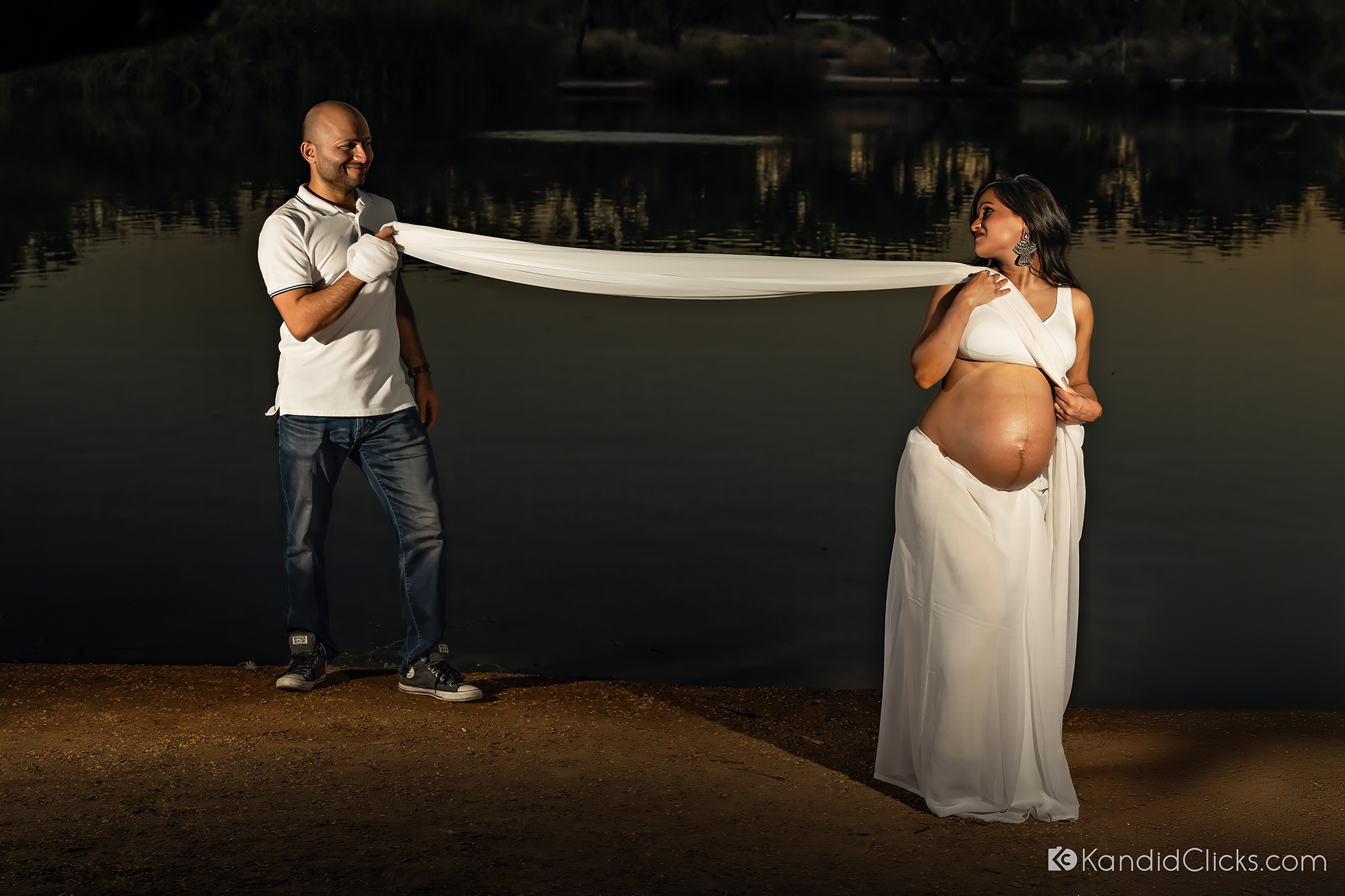 Maternity photographer captures joyful moment as couple poses by lake with playful fabric interaction