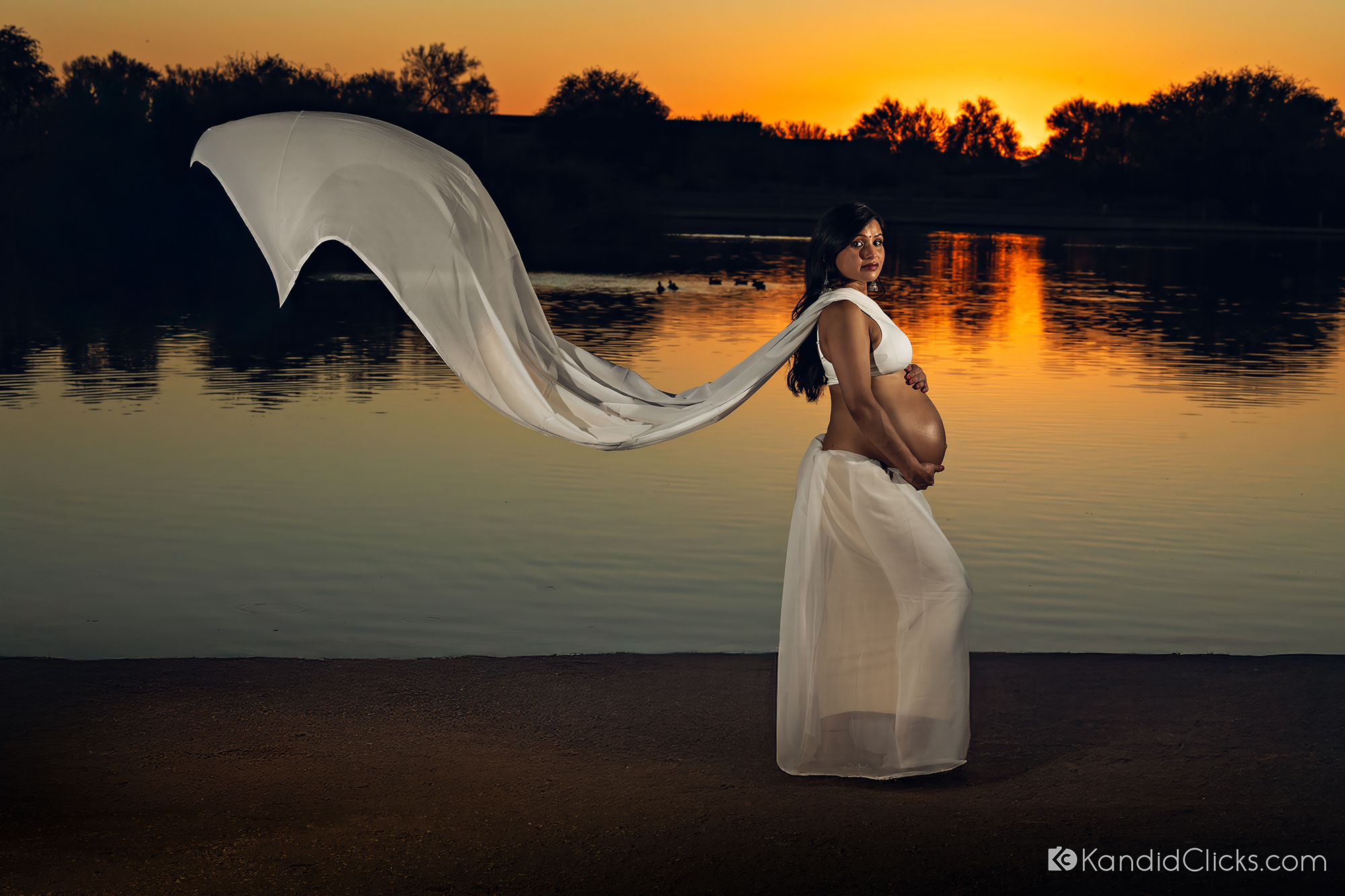 Maternity photographer captures elegant lakeside sunset portrait with flowing white fabric and soft reflections