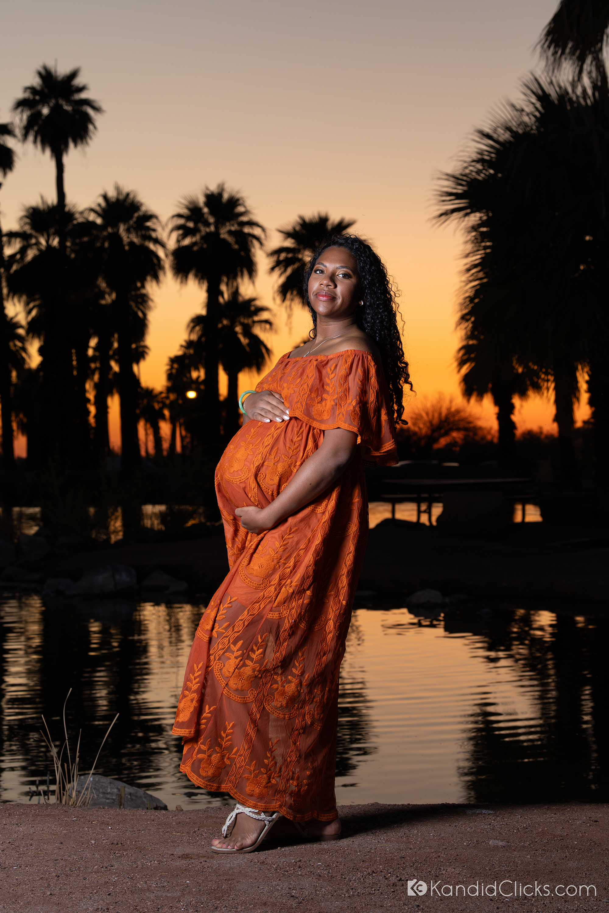 Maternity photographer captures glowing evening portrait of pregnant woman in orange dress by water and trees