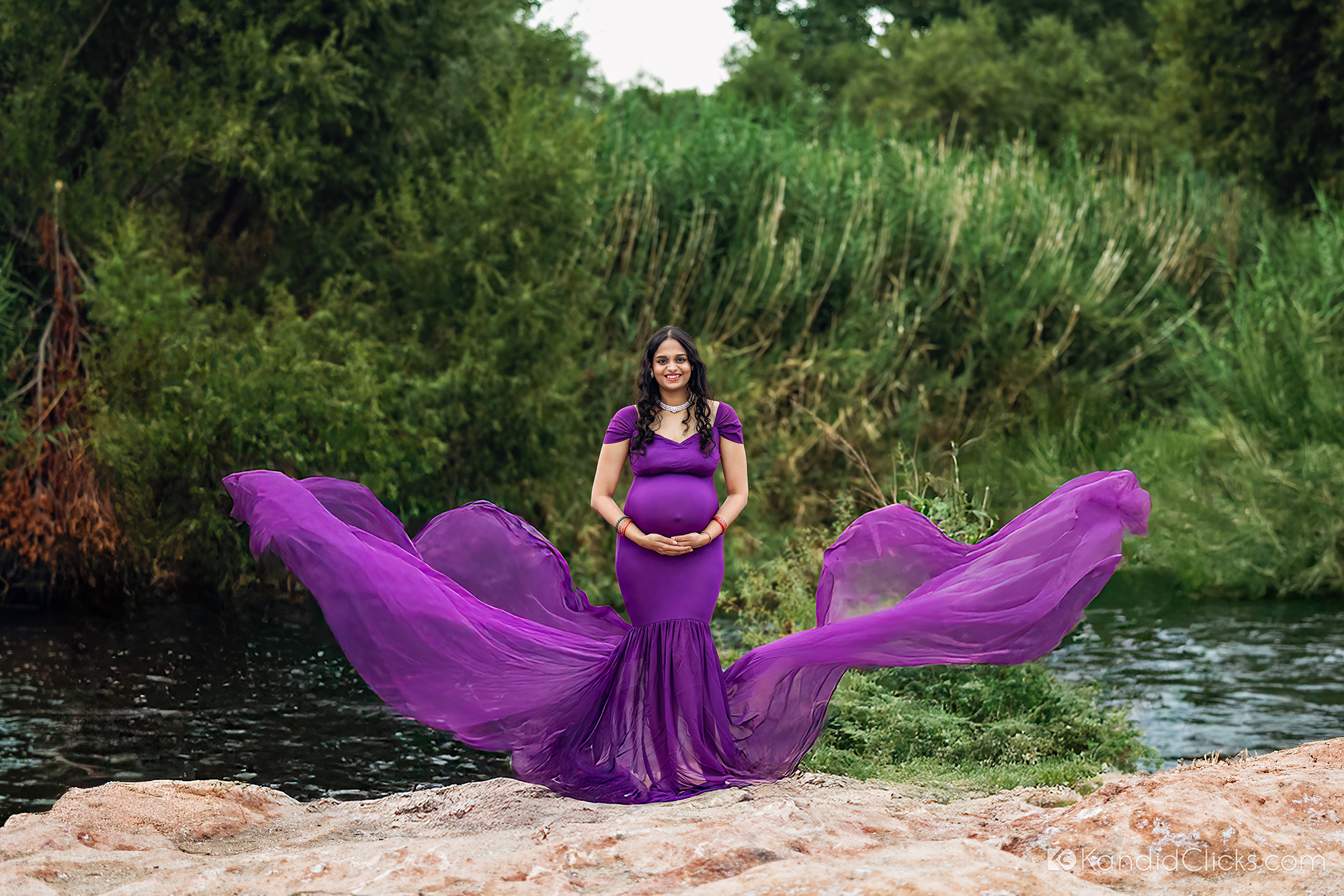 Pregnant woman poses confidently by a stream in a purple gown with extended flowing fabric in nature
