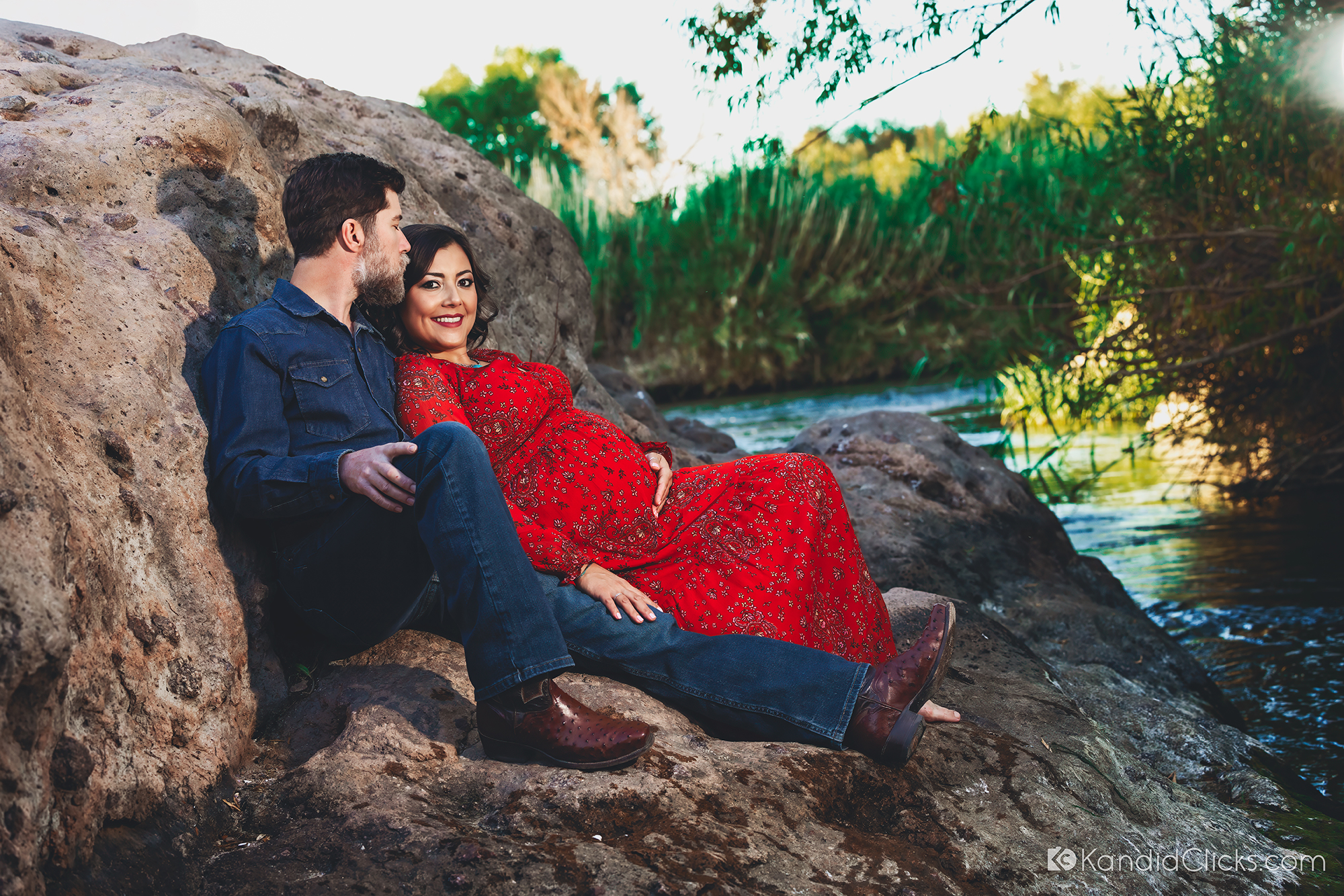 Pregnant woman in red floral dress leaning on her partner beside a scenic river during golden hour
