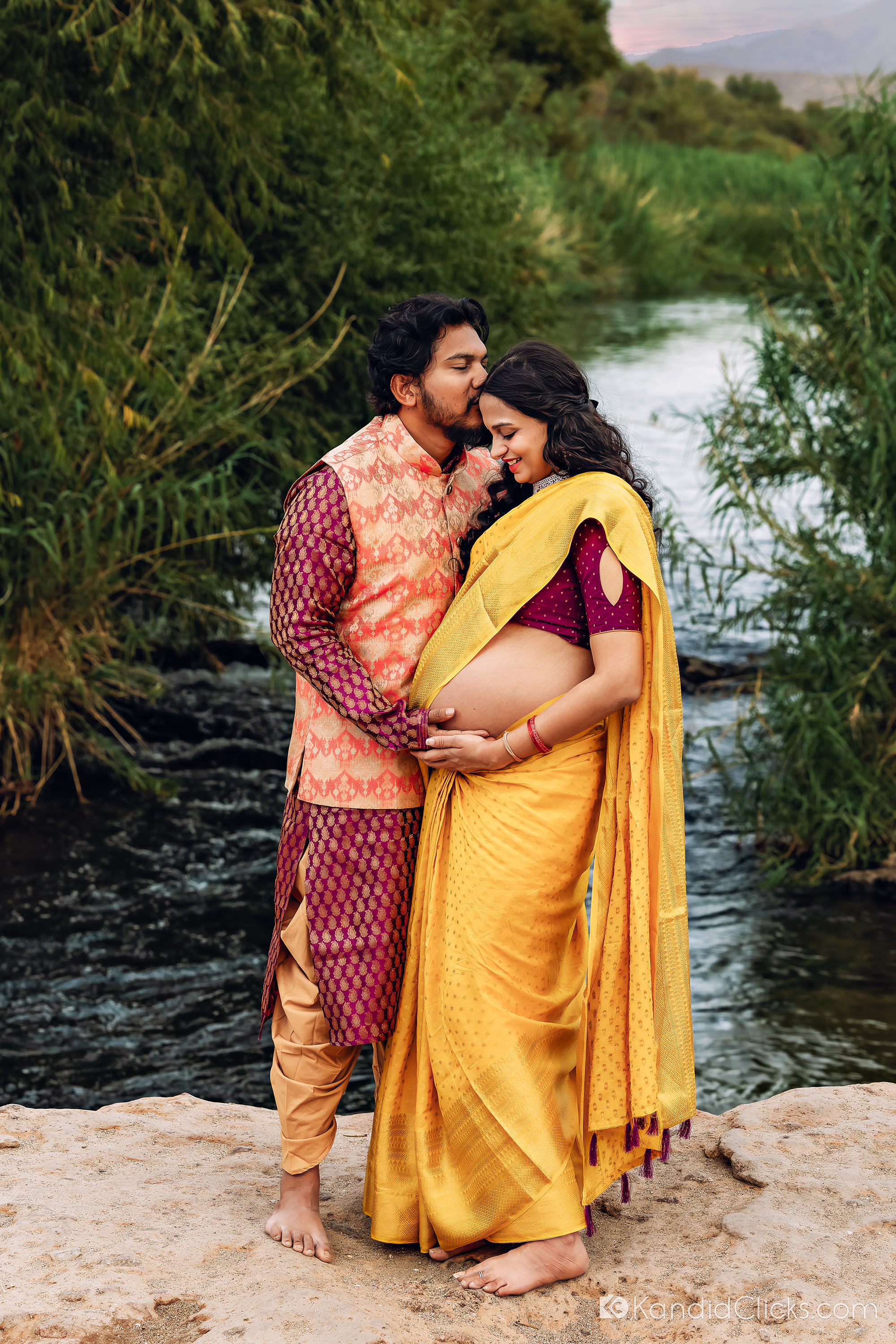 Pregnant woman in yellow saree stands with her partner as they share a quiet embrace near the river