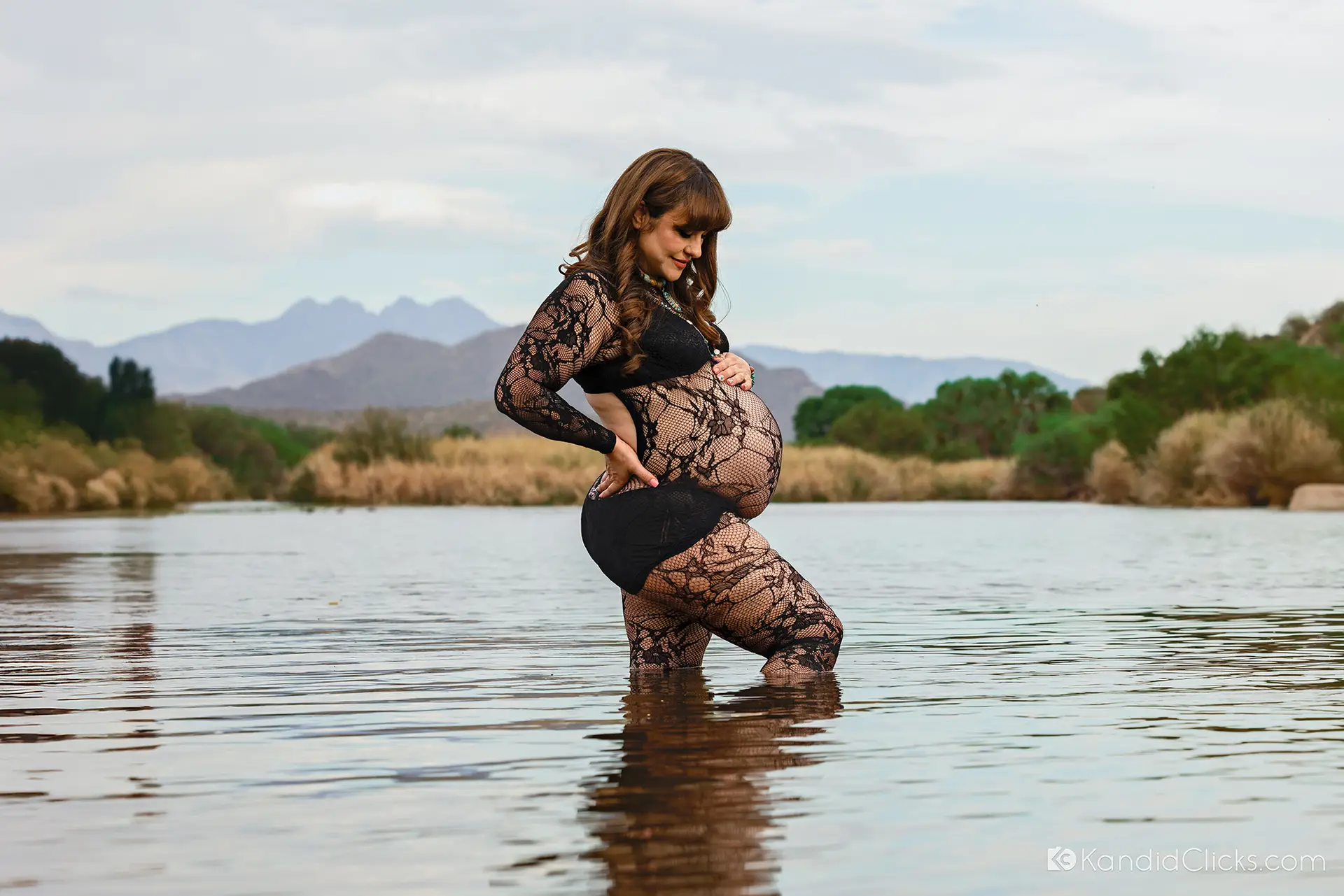 Maternity photographer captures pregnant woman in black lace outfit standing in water with scenic backdrop