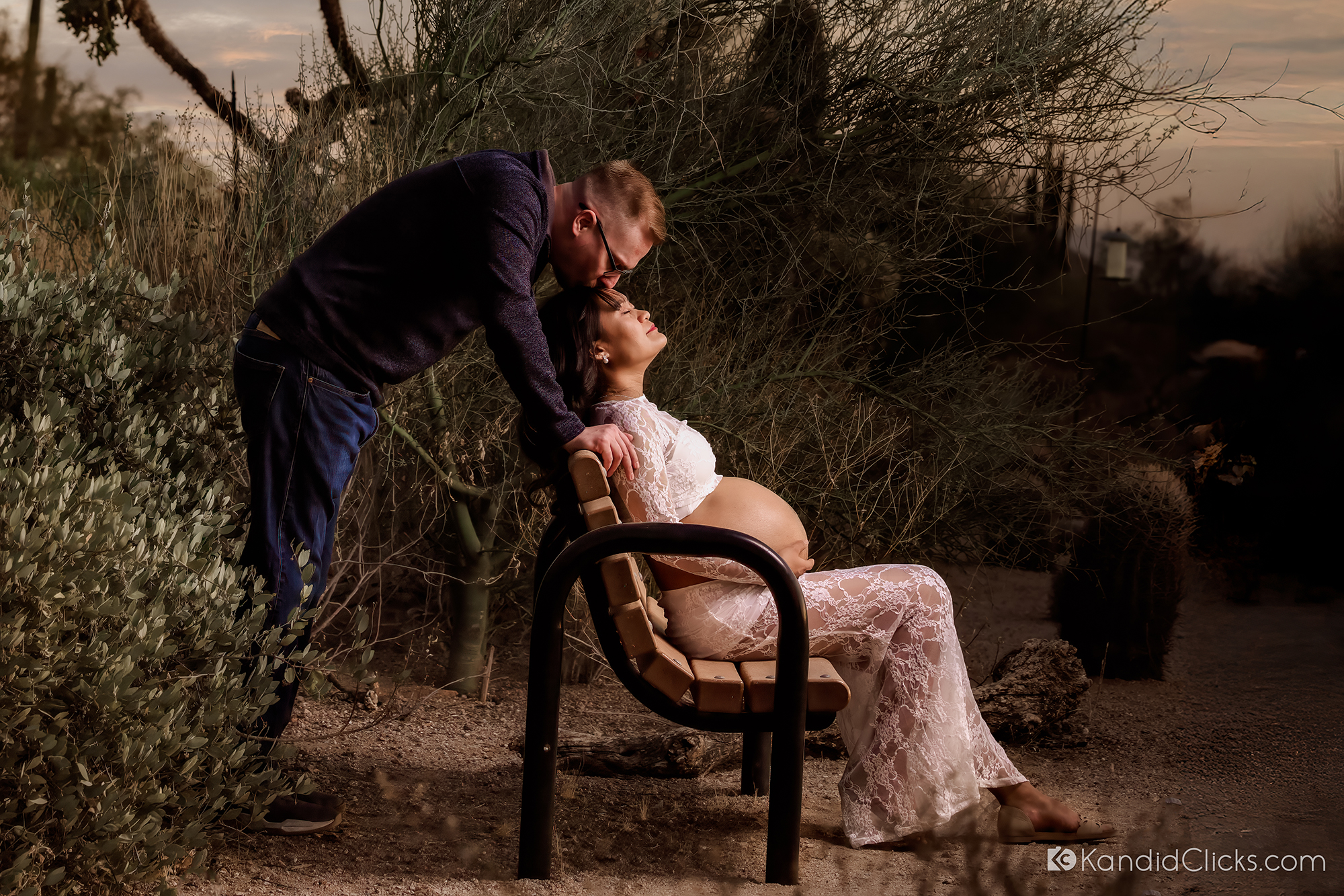 Man kisses pregnant woman's forehead while she relaxes on a park bench during maternity photoshoot
