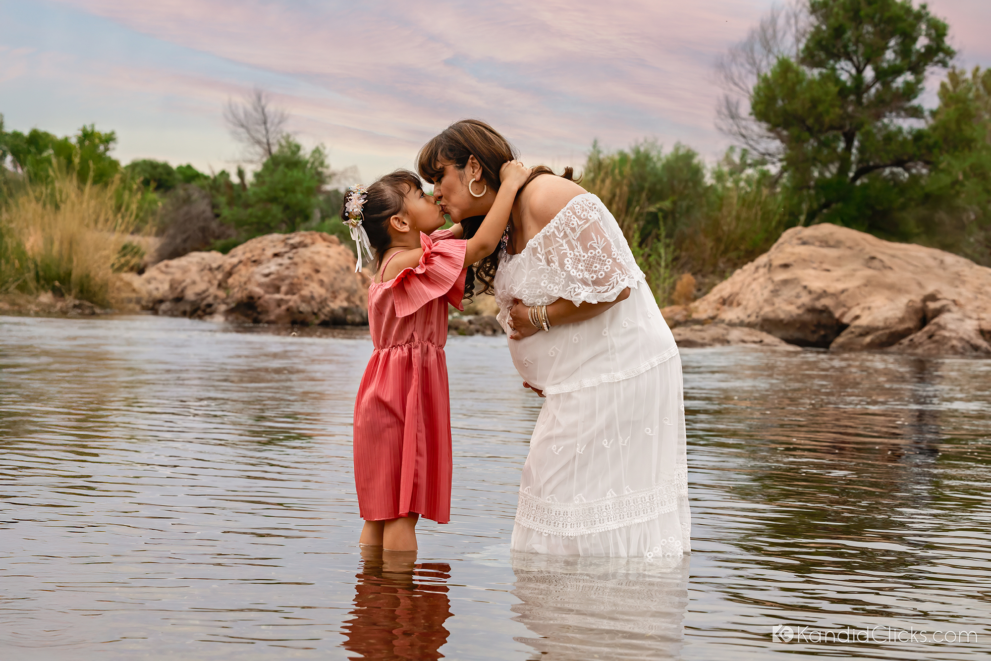 Maternity photographer captures mom-to-be sharing a loving kiss with her daughter in a peaceful river setting