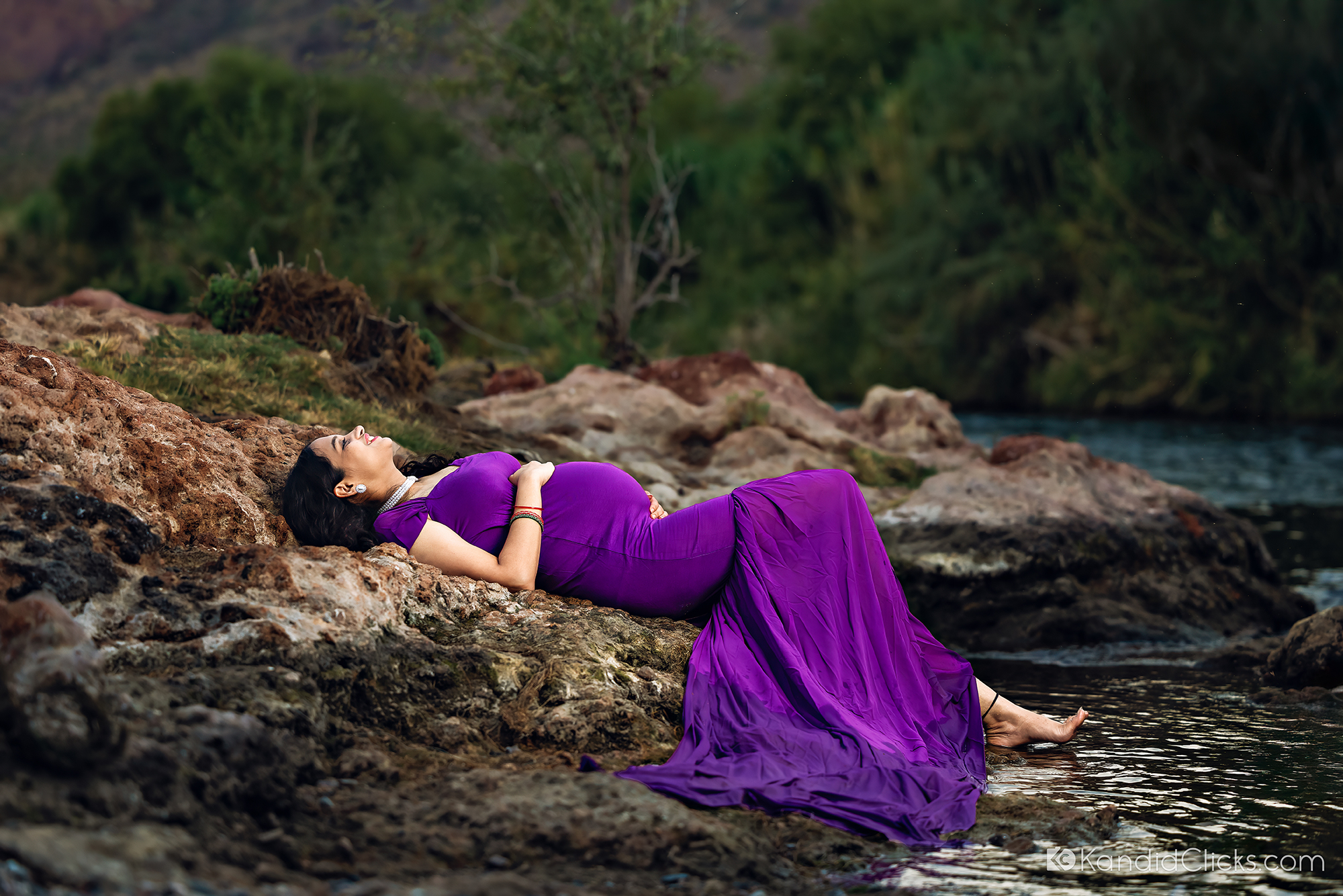Maternity photographer captures serene portrait of expecting mom lying on rocks in a purple dress by the river