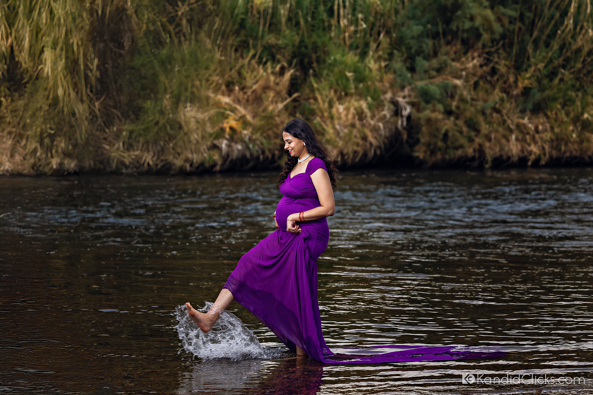 Maternity photographer captures playful moment of pregnant woman splashing water in purple dress by the river