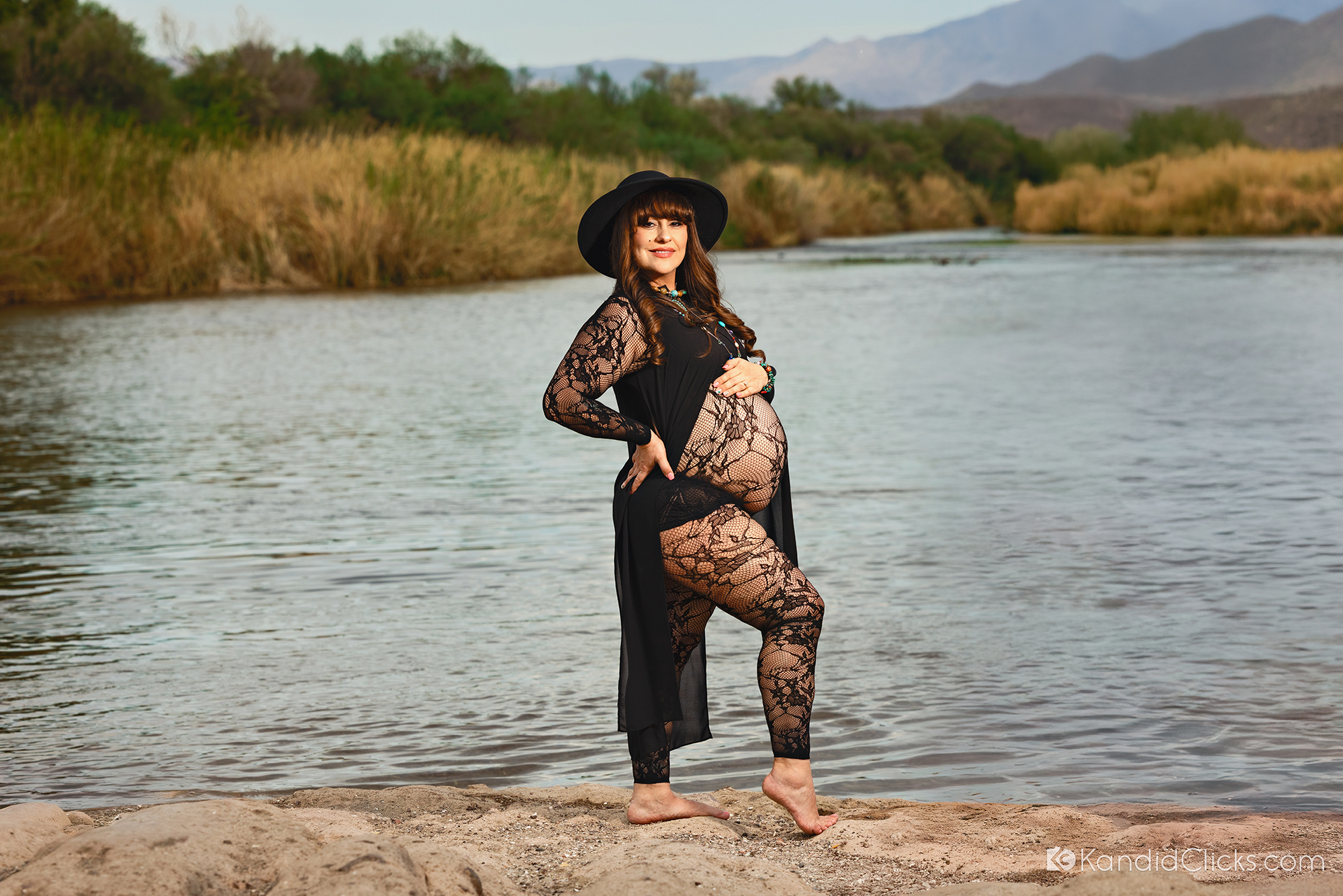 Maternity photographer captures barefoot expecting mother in lace dress by a calm river and mountain view