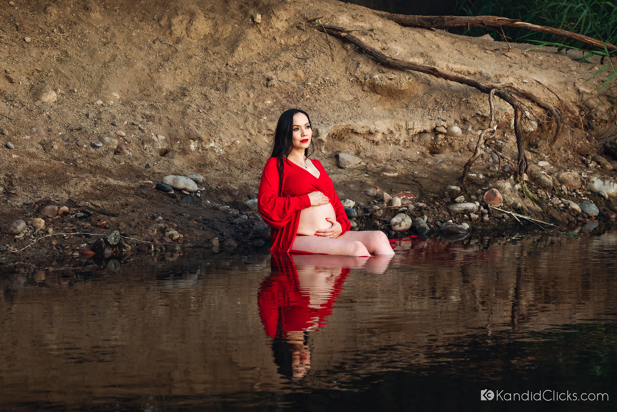 Pregnant woman in red gown sitting peacefully in a shallow river during a maternity photoshoot