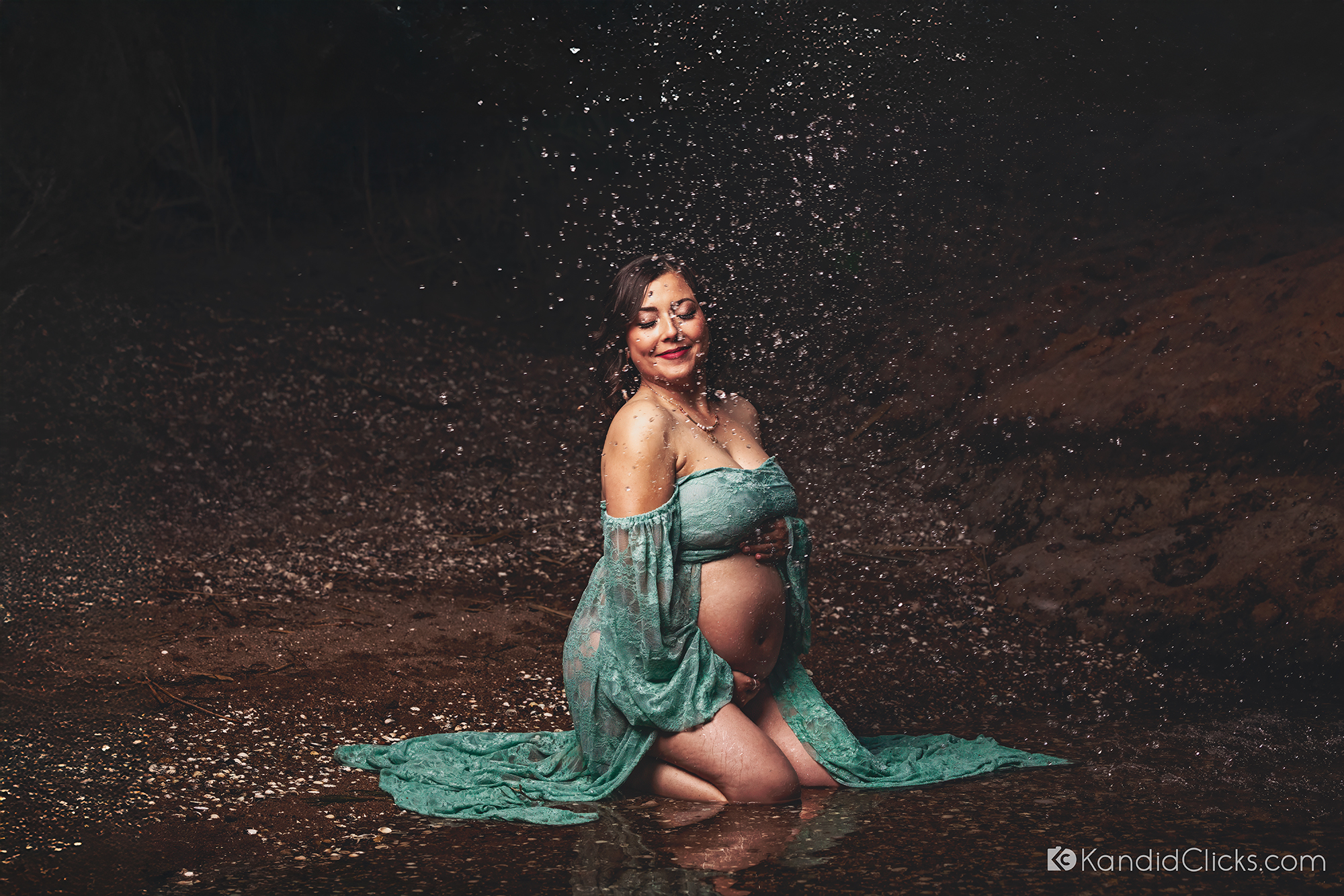 Pregnant woman in green lace gown enjoying a splash of water during artistic maternity shoot