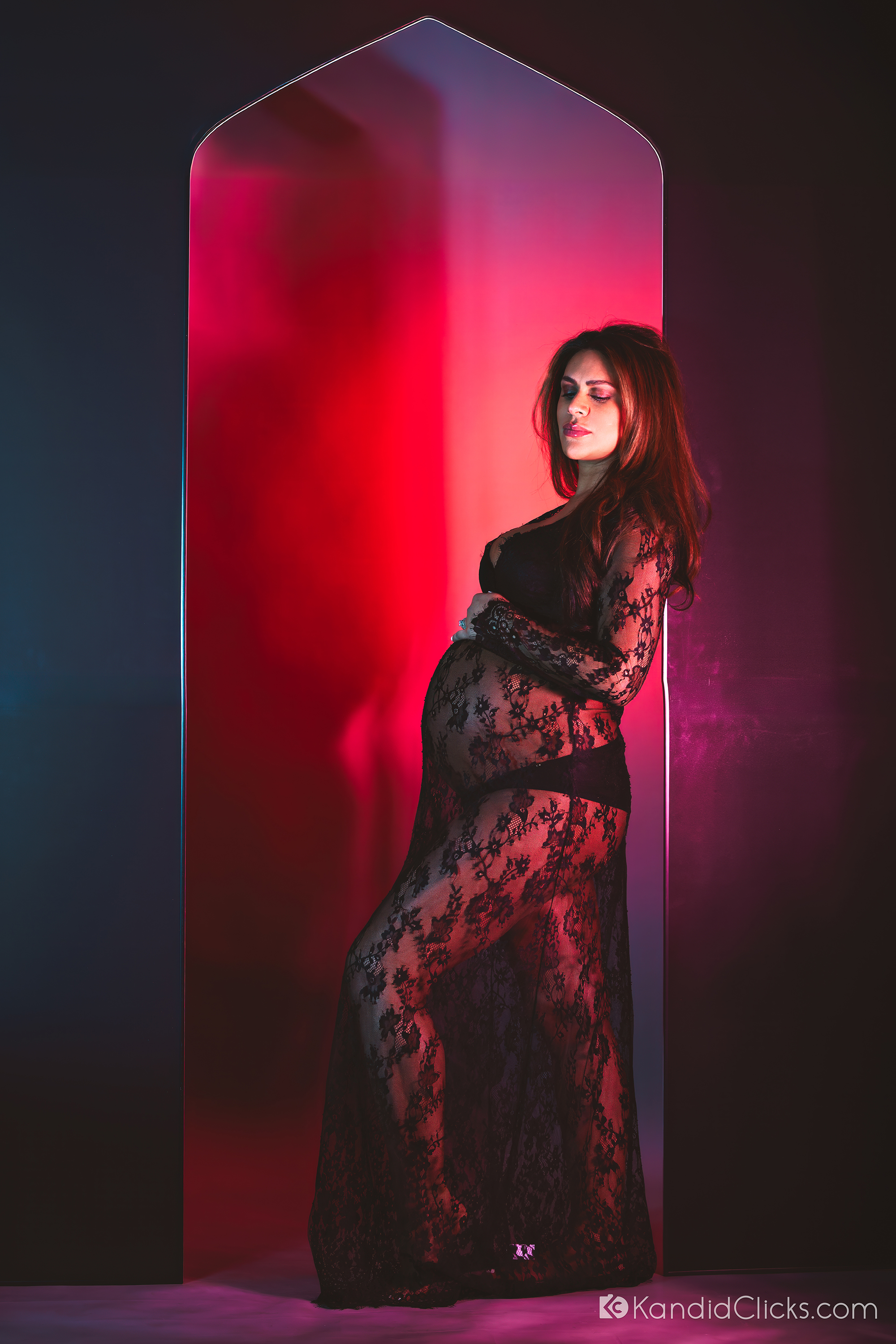 Pregnant woman in sheer black lace gown leaning against red-lit arch in creative studio setting