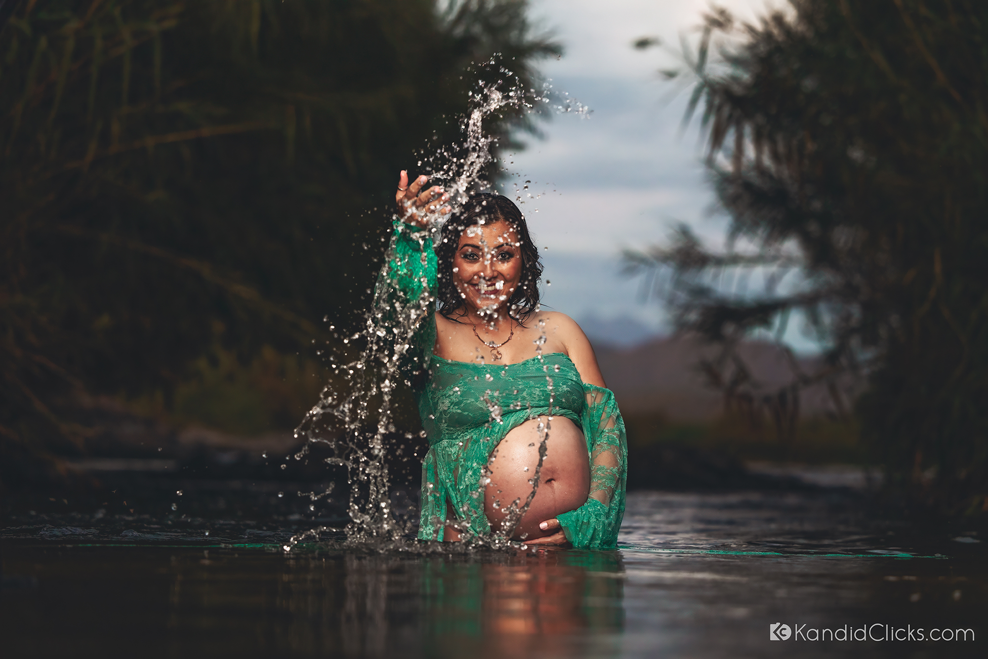 Pregnant woman in green lace dress splashing water during river maternity photoshoot