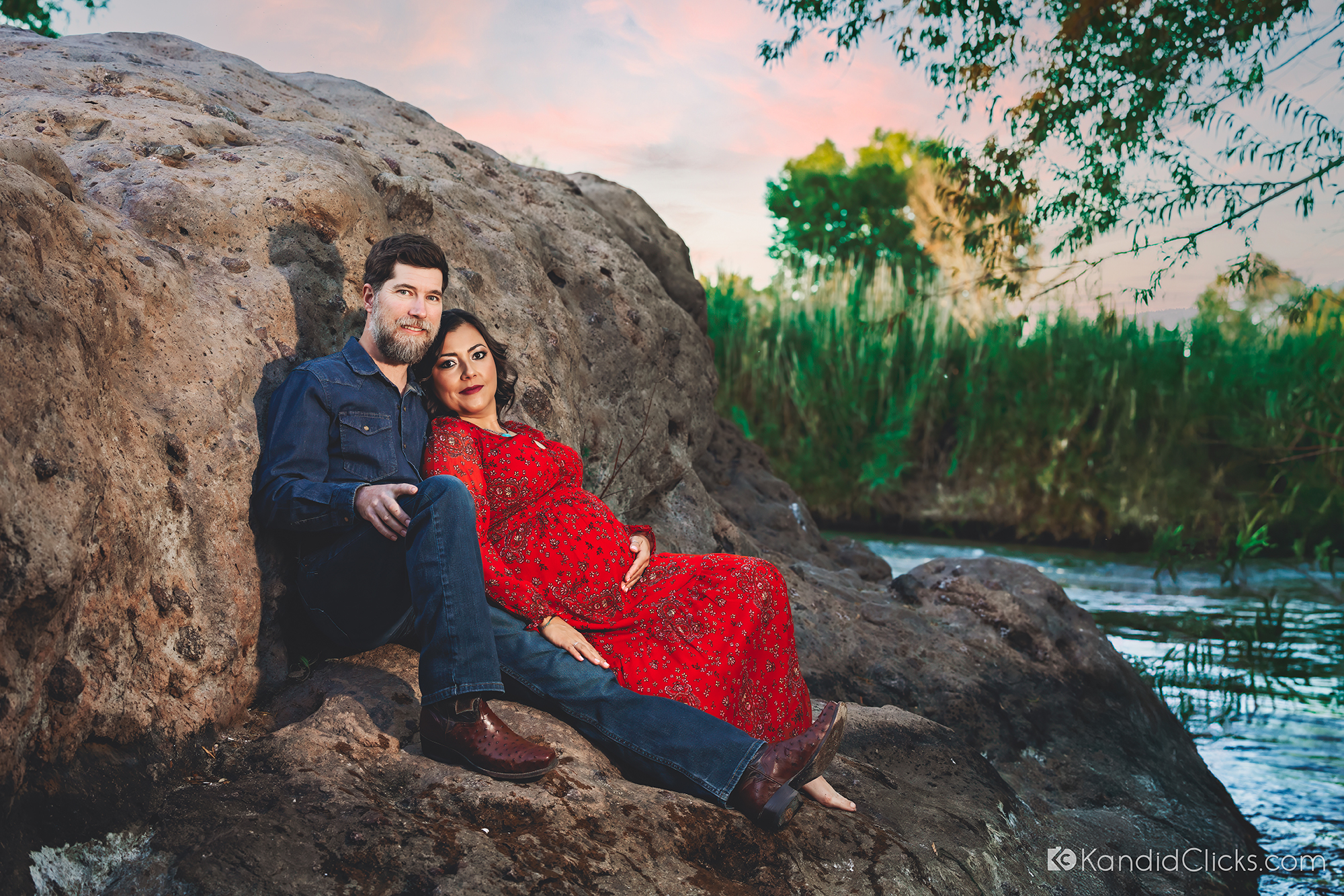 Pregnant woman in red dress leaning on partner by the river during maternity shoot