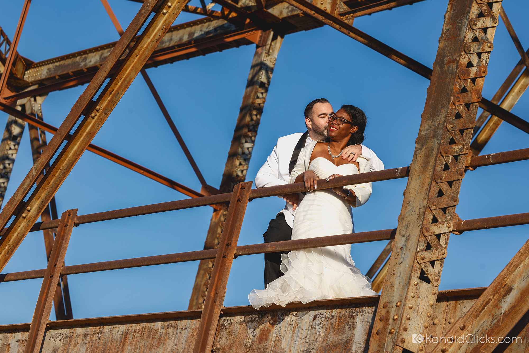Bride and groom embracing on a rusted iron bridge against a clear blue sky.