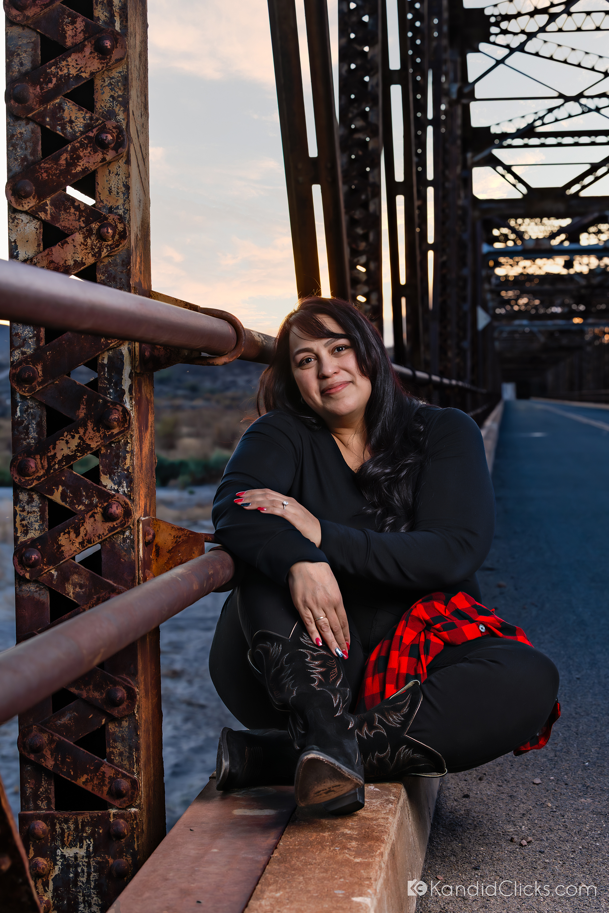Woman in black outfit and cowboy boots sitting on a rustic bridge at sunset.