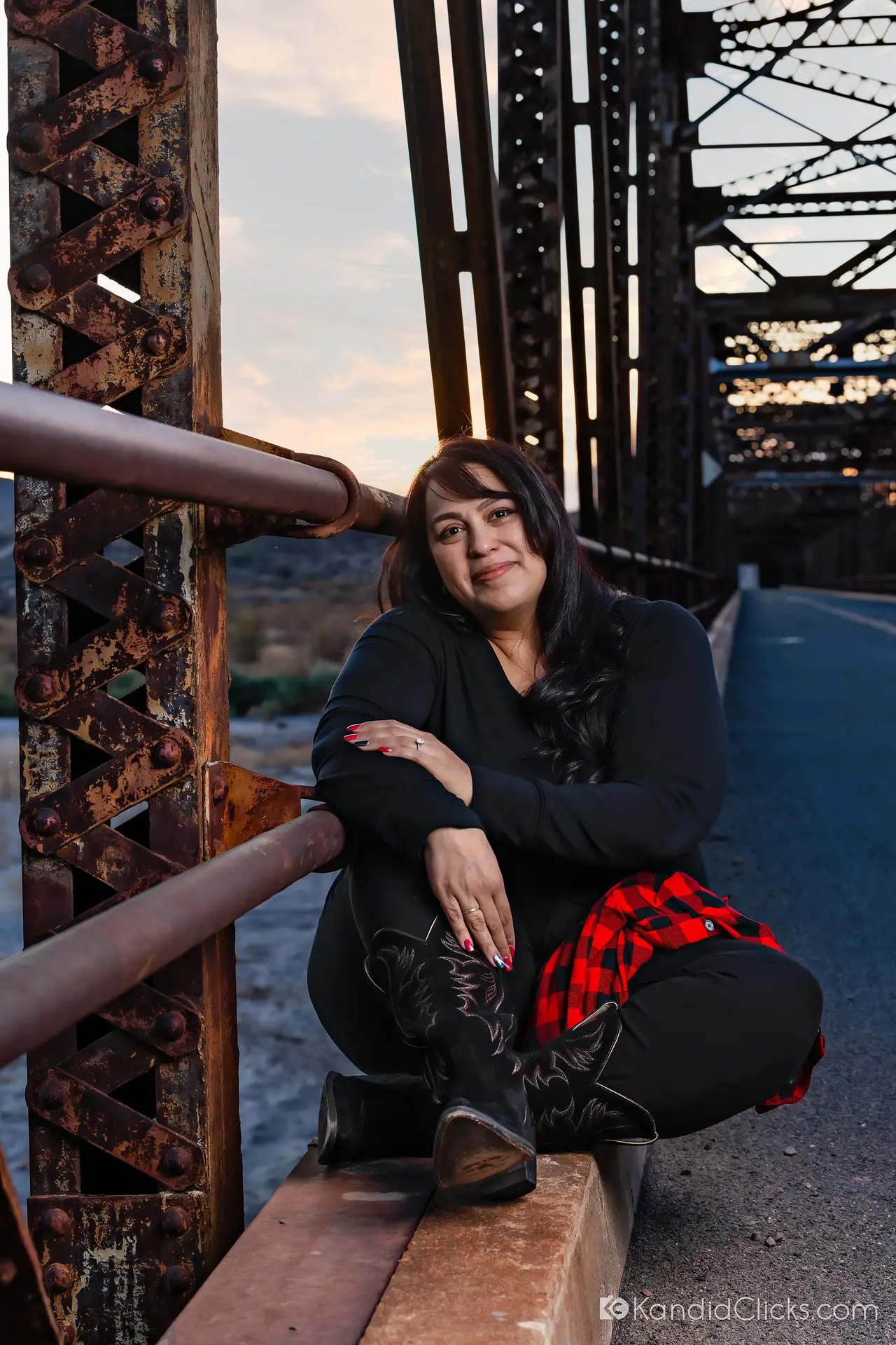 Woman in black outfit and cowboy boots sitting on a rustic bridge at sunset.