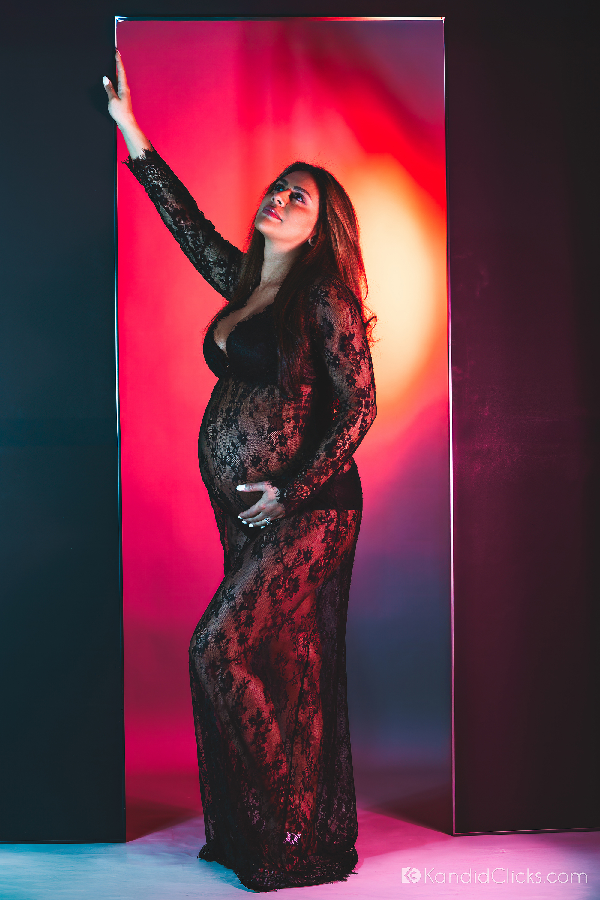 Pregnant woman in sheer black lace dress posing confidently in dramatic colored studio lighting