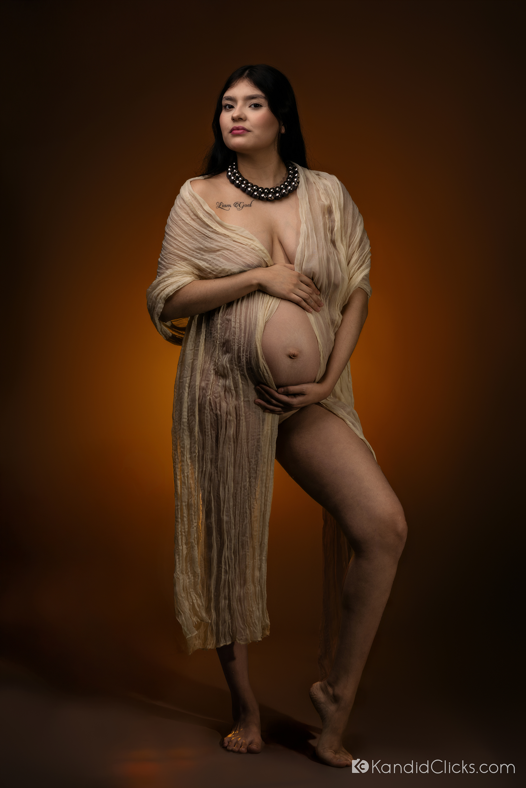 Pregnant woman in sheer cream gown posing confidently in a studio maternity shoot with dramatic warm lighting