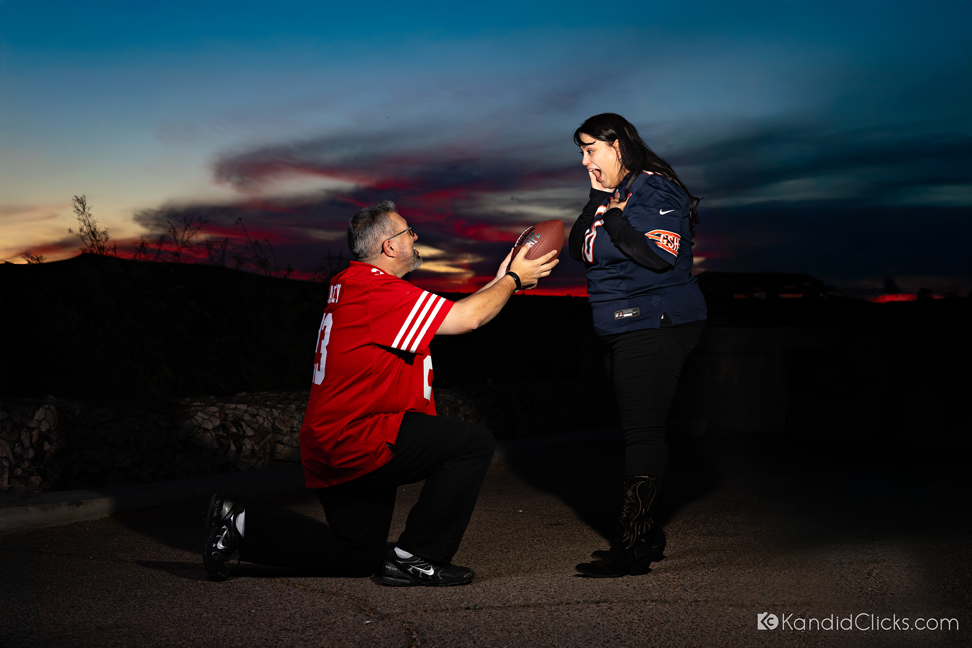 Man proposing with a football at sunset while woman reacts emotionally.