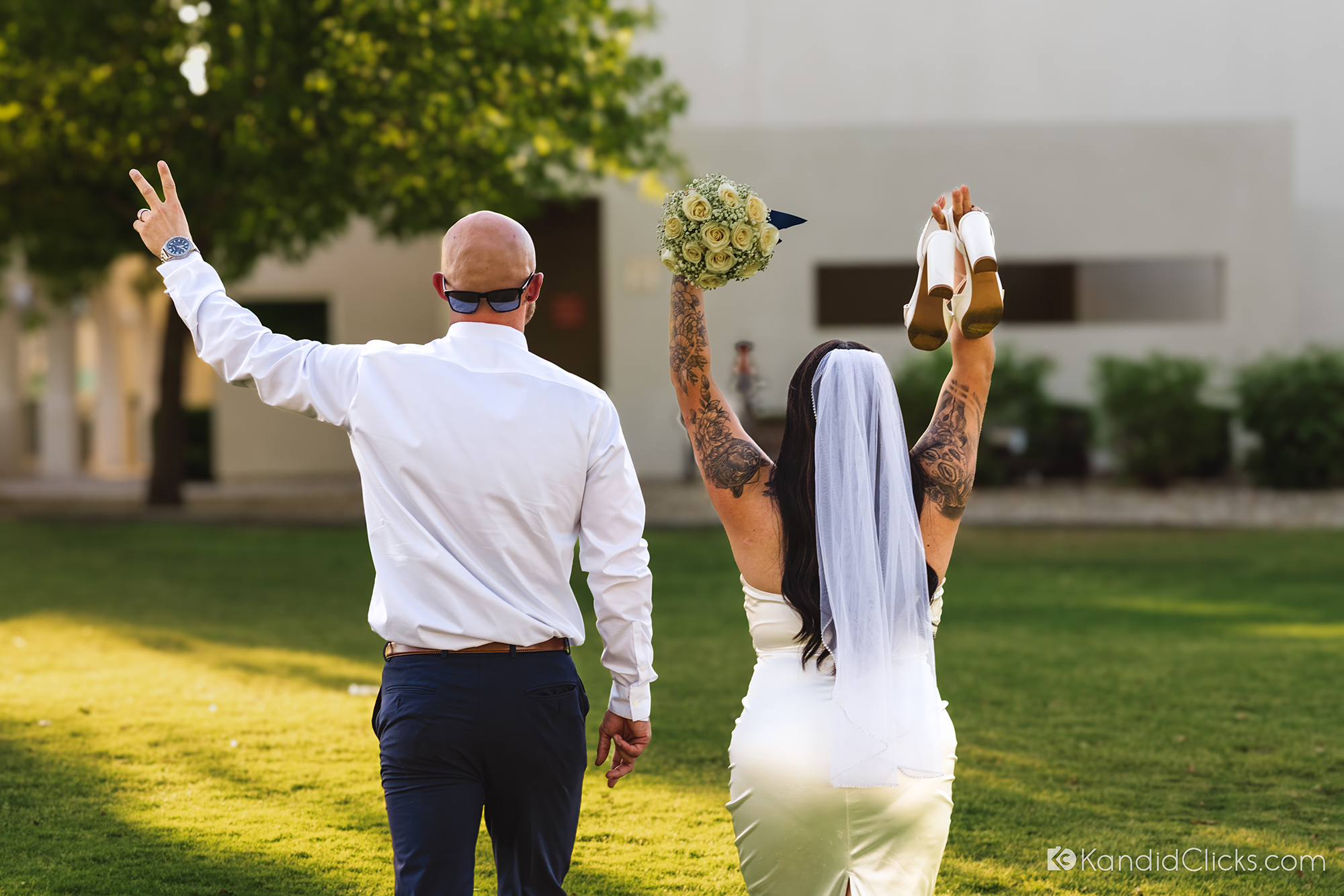 Bride and groom walk away with hands raised in joy, holding bouquet and shoes on their Arizona wedding day