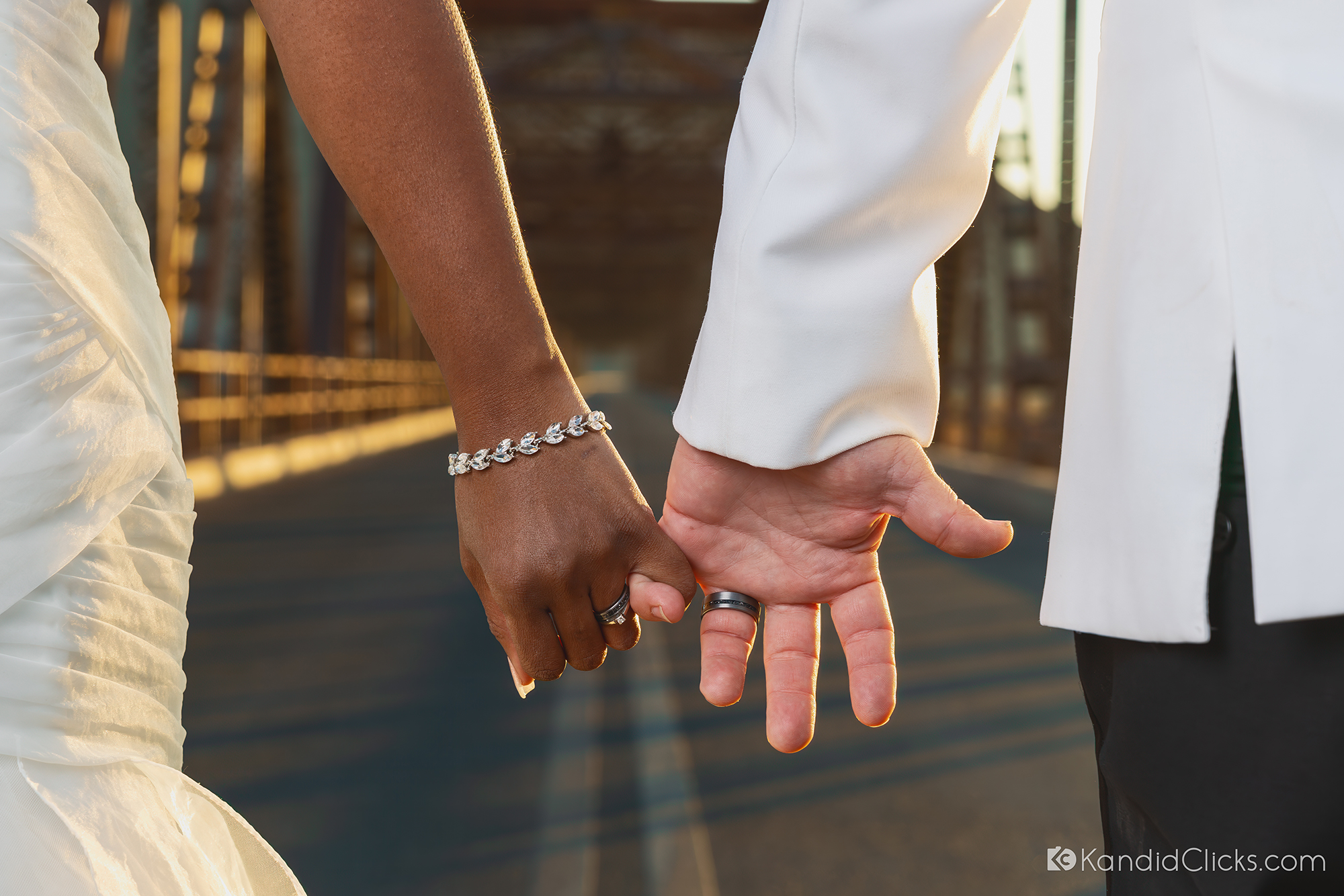 Bride and groom holding hands with wedding rings visible at golden hour on a rustic bridge
