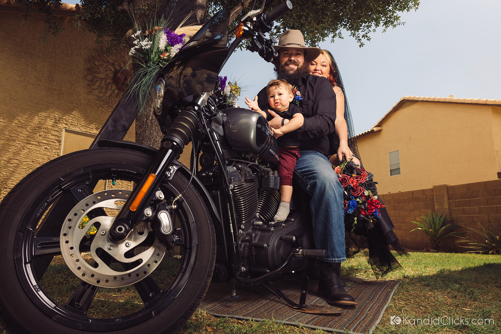 Bride, groom, and toddler pose together on a Harley-Davidson during their outdoor wedding