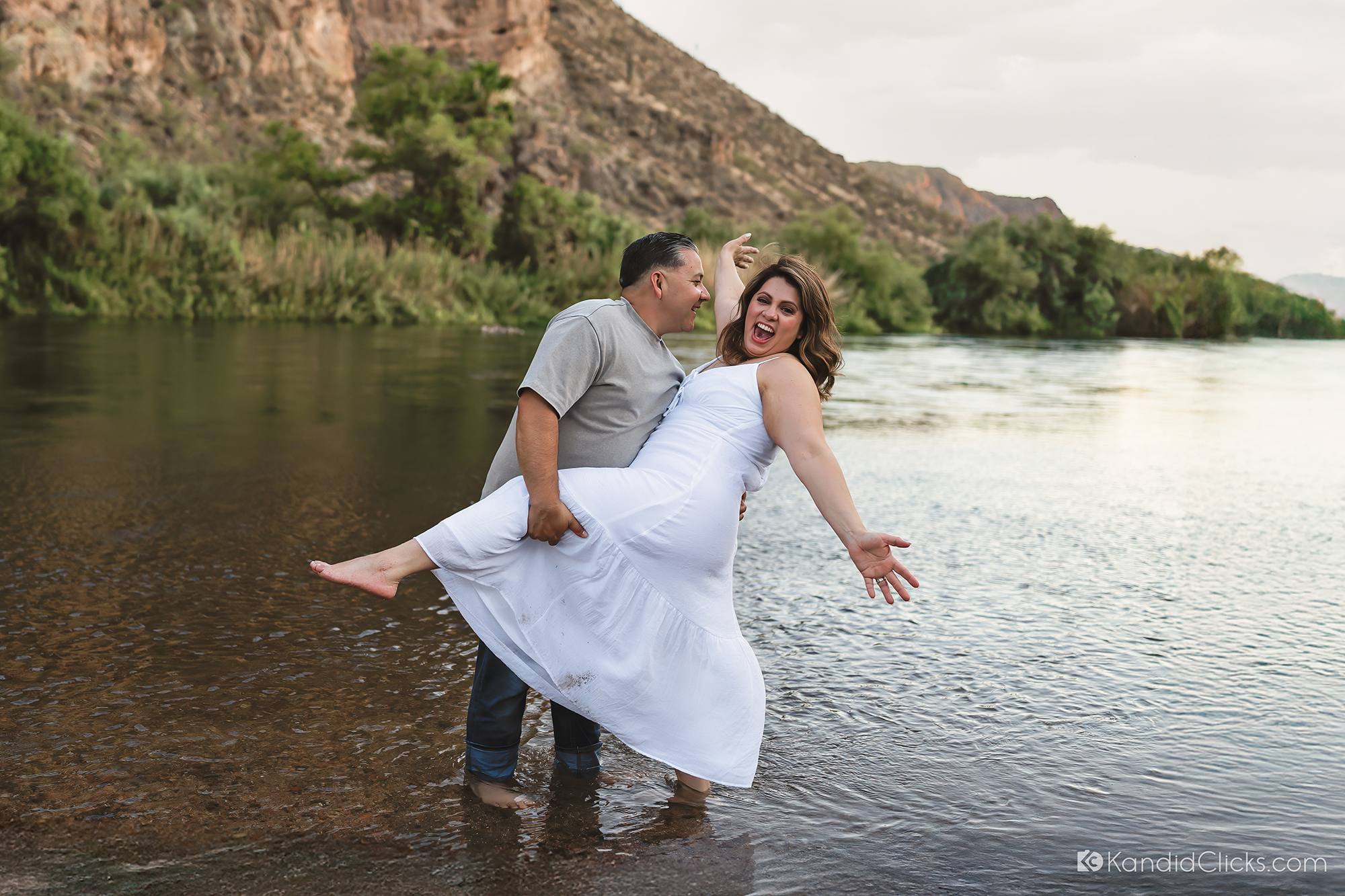 Man lifting woman playfully in river with mountains and greenery in the background
