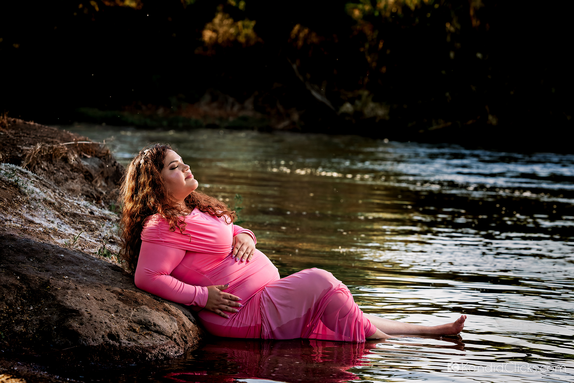 Pregnant woman in pink gown reclining by riverbank during peaceful maternity photoshoot.