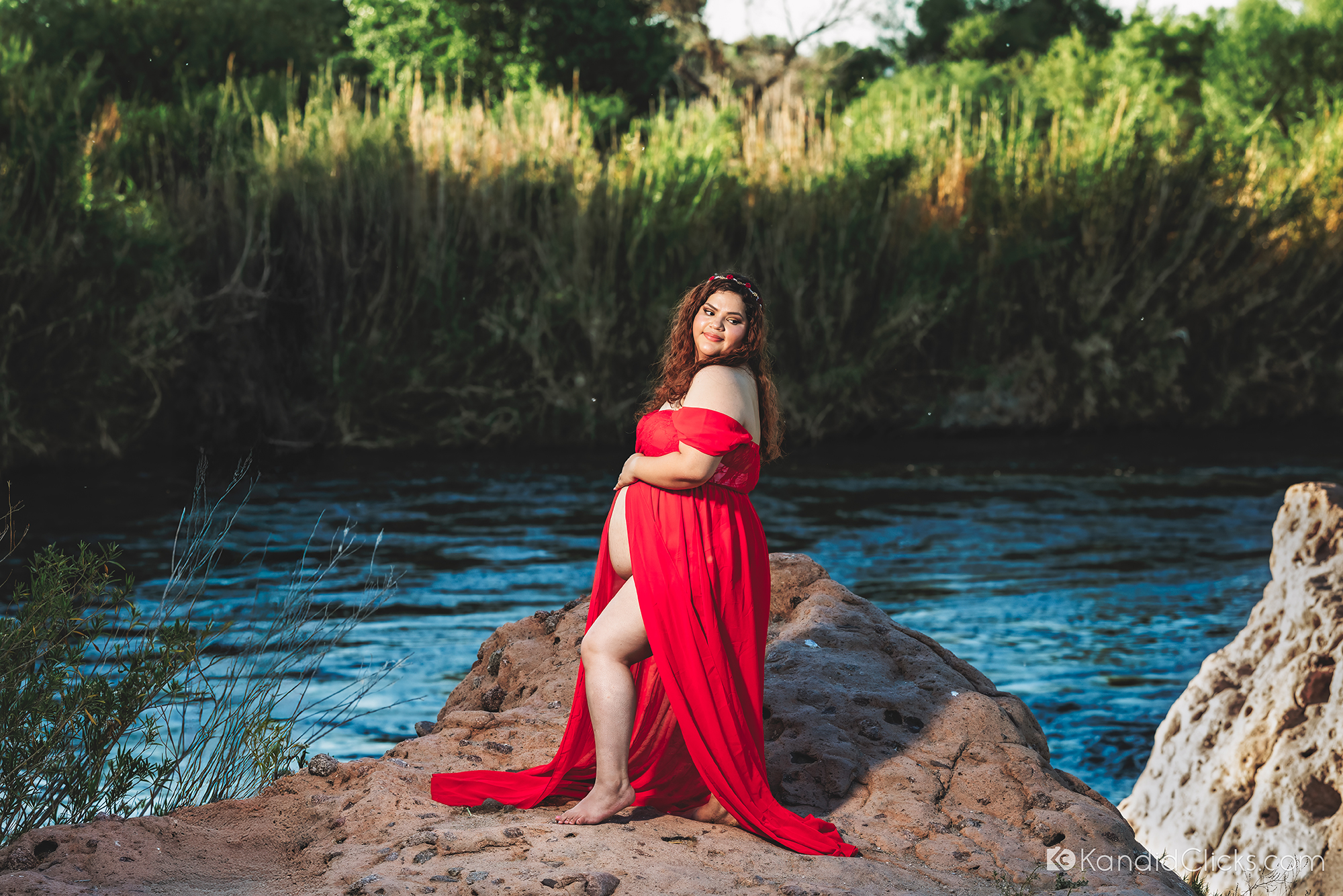 Pregnant woman in red gown standing confidently on river rocks during golden hour maternity shoot.