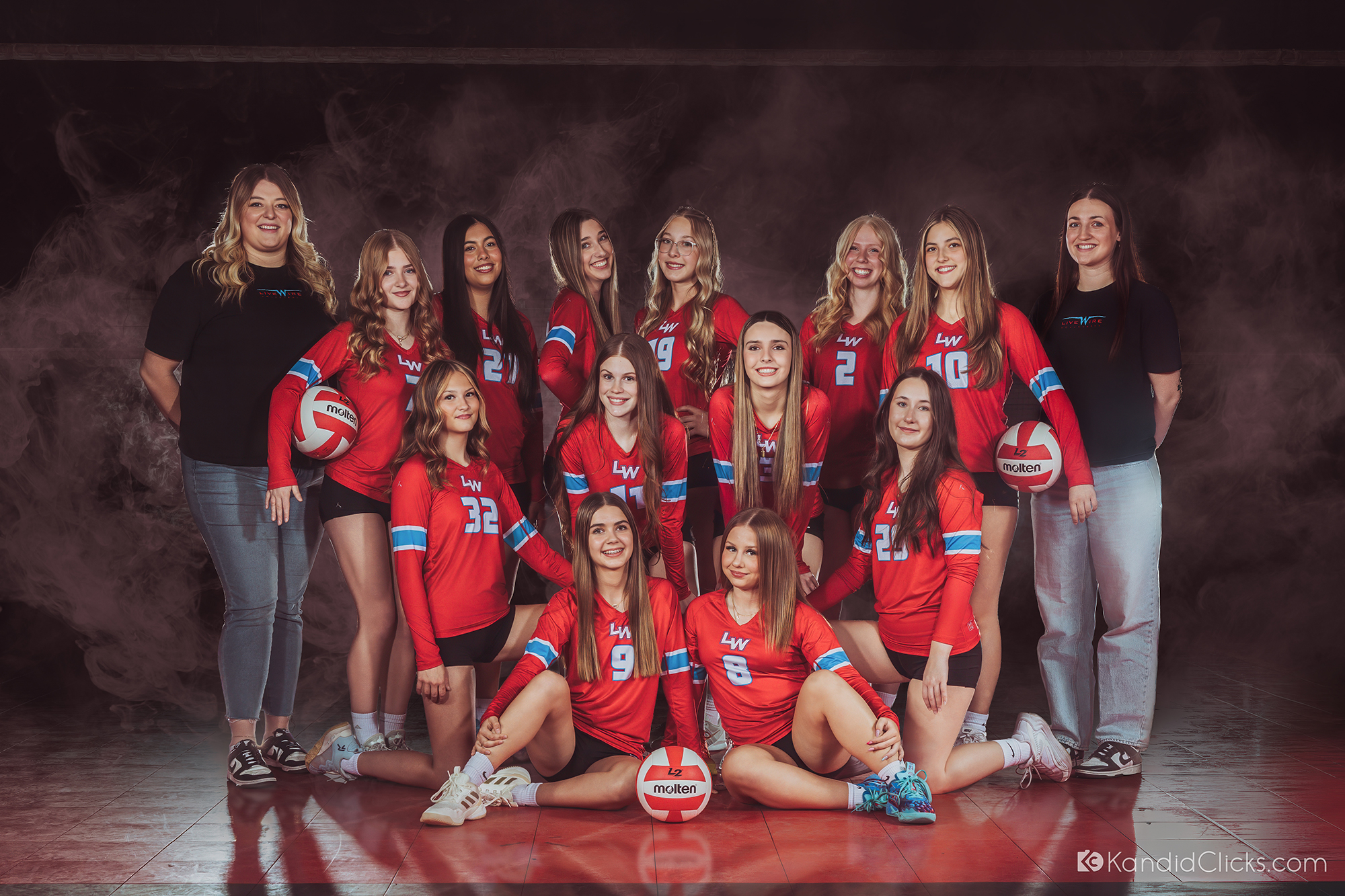 A beautifully lit group portrait of a female volleyball squad and their two coaches smiling on a smoky red court, representing striking athlete portraits.