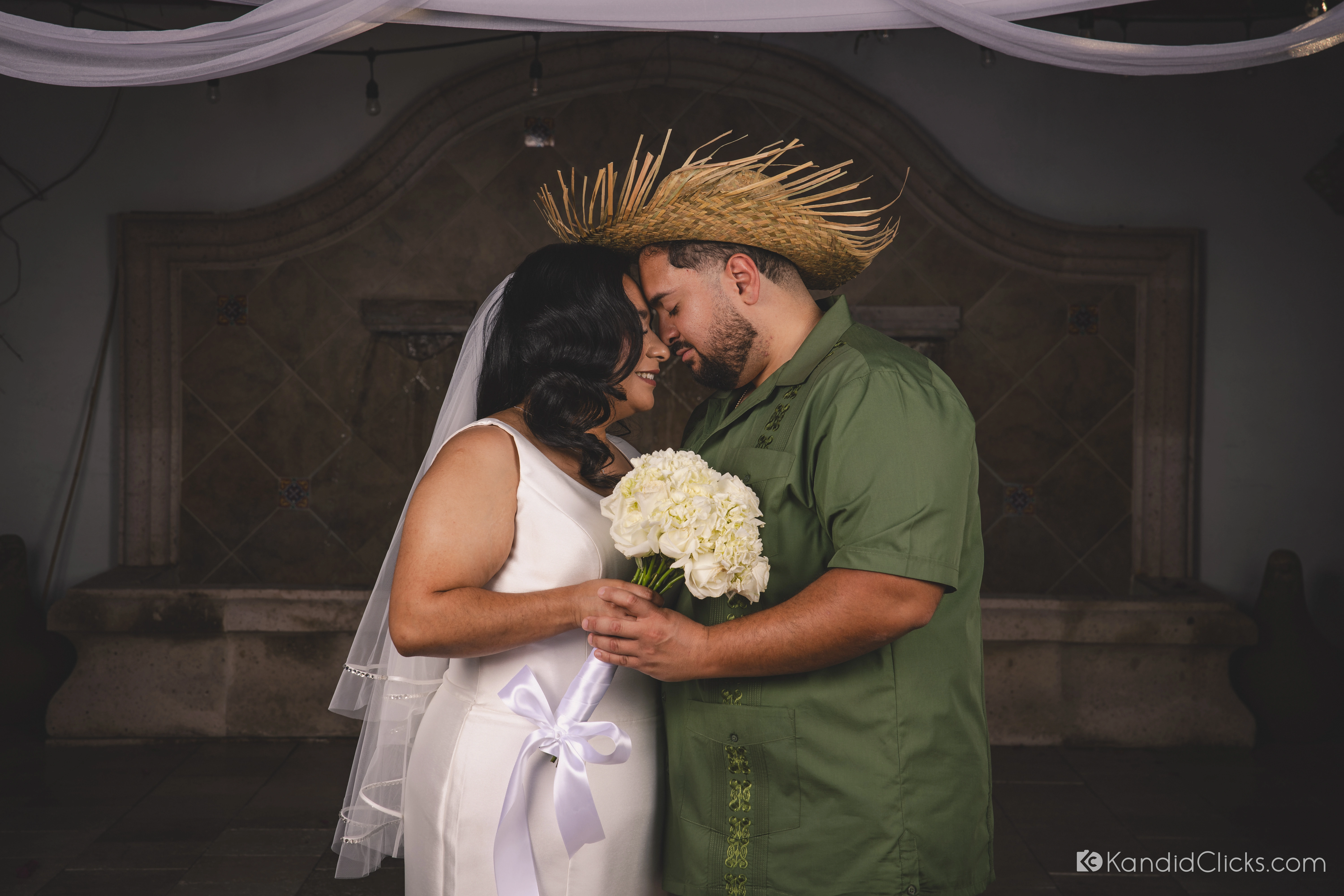 Bride Alba and groom Lucas resting forehead to forehead, the groom in a straw hat, against a moody fountain backdrop at their Phoenix wedding.